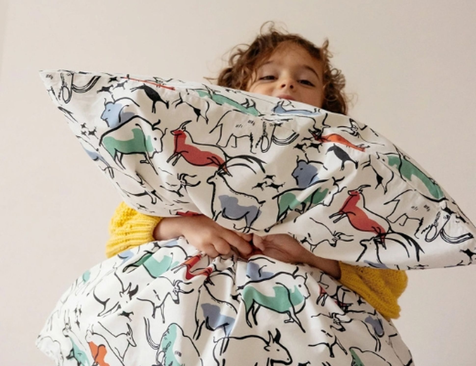 Enfant tenant un oreiller avec des illustrations colorées de chevaux, vêtu d'un pull jaune, debout devant un fond uni.