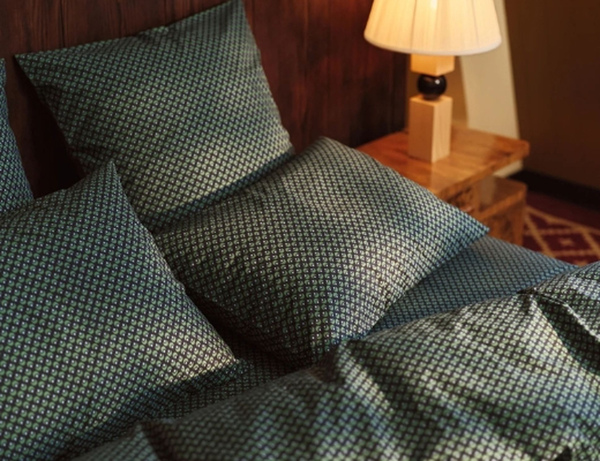 Un lit orné d'oreillers et d'une couette à motifs verts, à côté d'une table de chevet en bois sur laquelle repose une lampe, adossé à un mur lambrissé.
