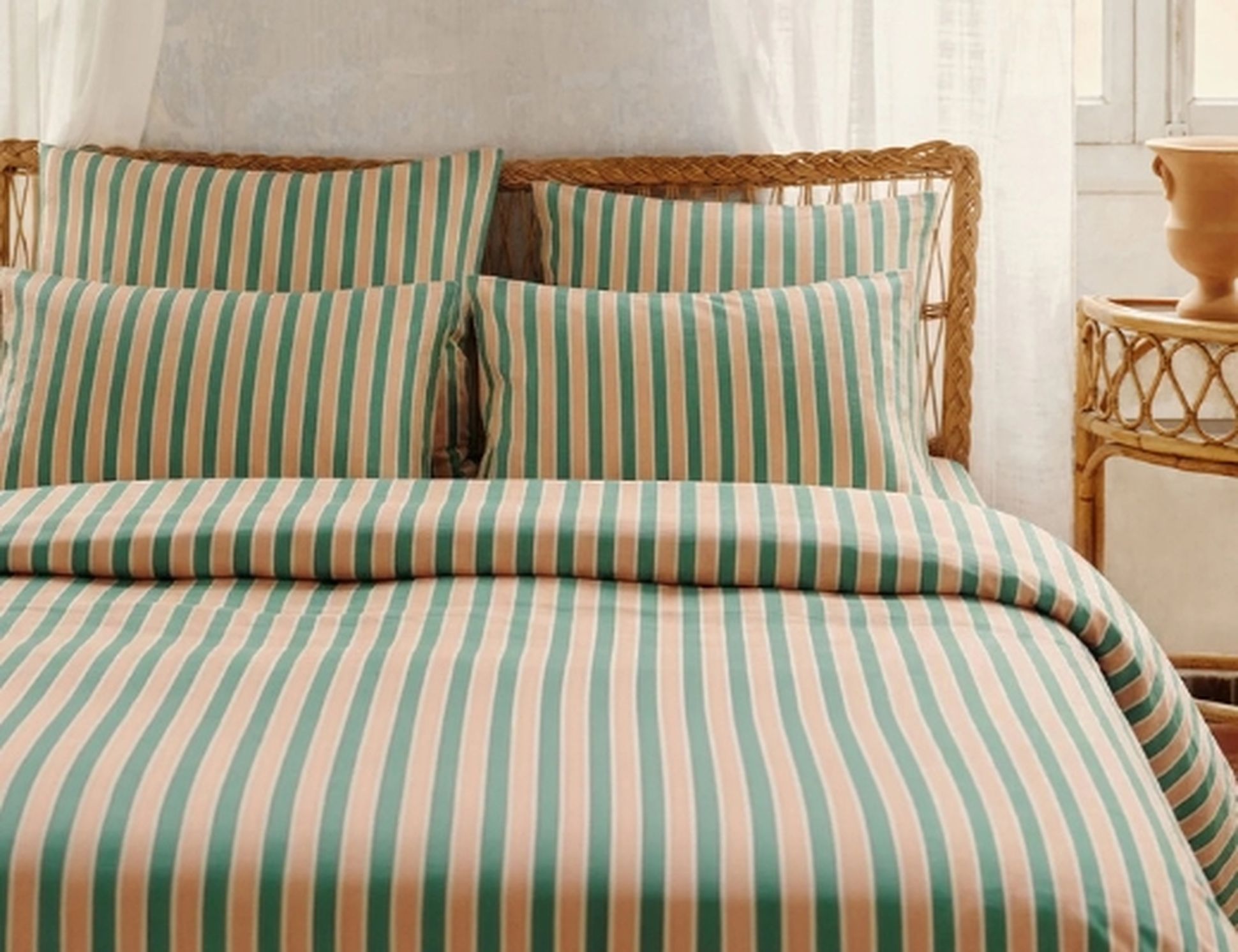 Ensemble de literie rayé dans les tons verts, roses et blancs, posé sur un lit en rotin. La lumière filtre à travers les rideaux et se pose sur une table de chevet en osier.