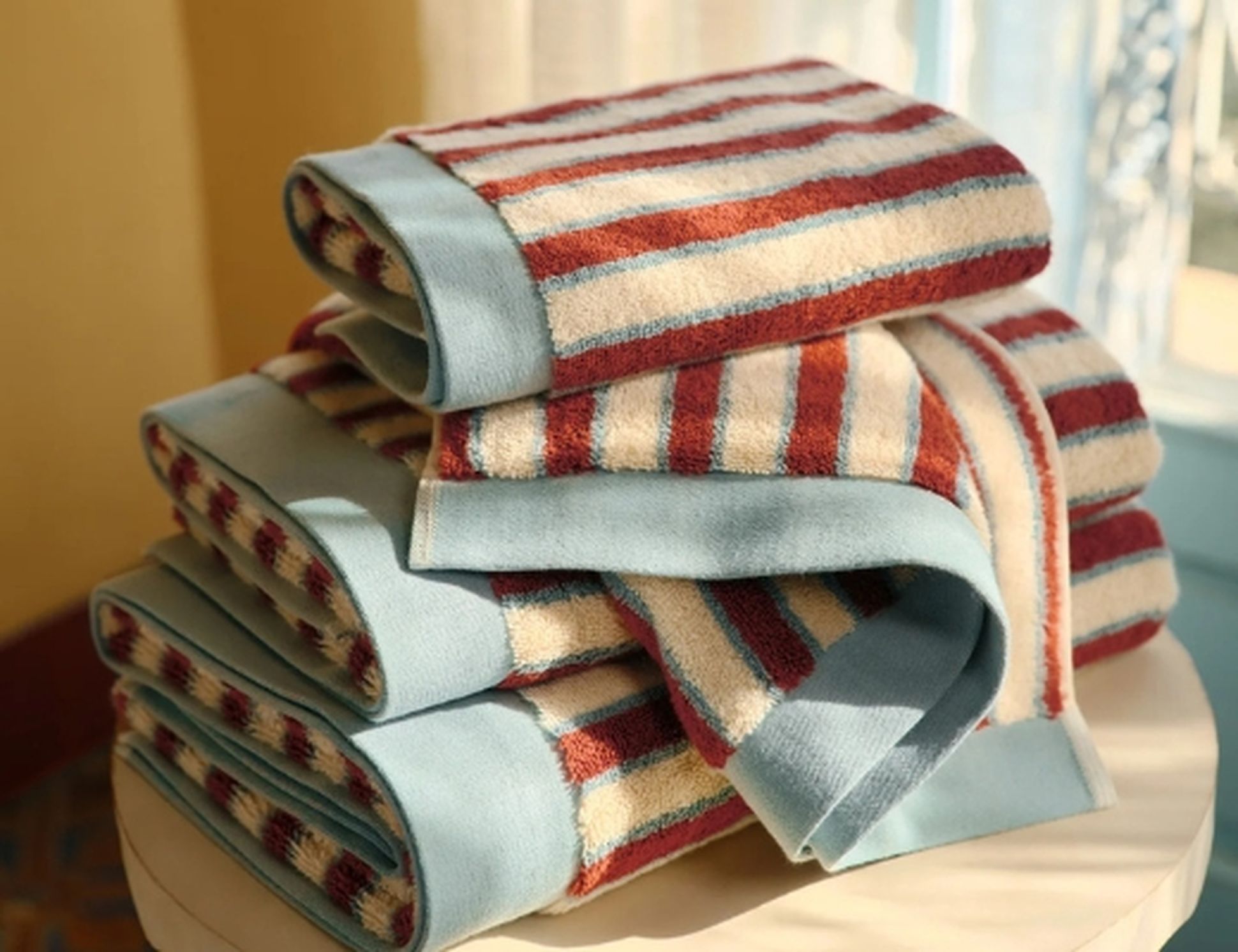 Une pile de serviettes pliées, rayées de rouge, de bleu et de beige, posée sur une table claire, baignée par la lumière chaude du soleil près d'une fenêtre.