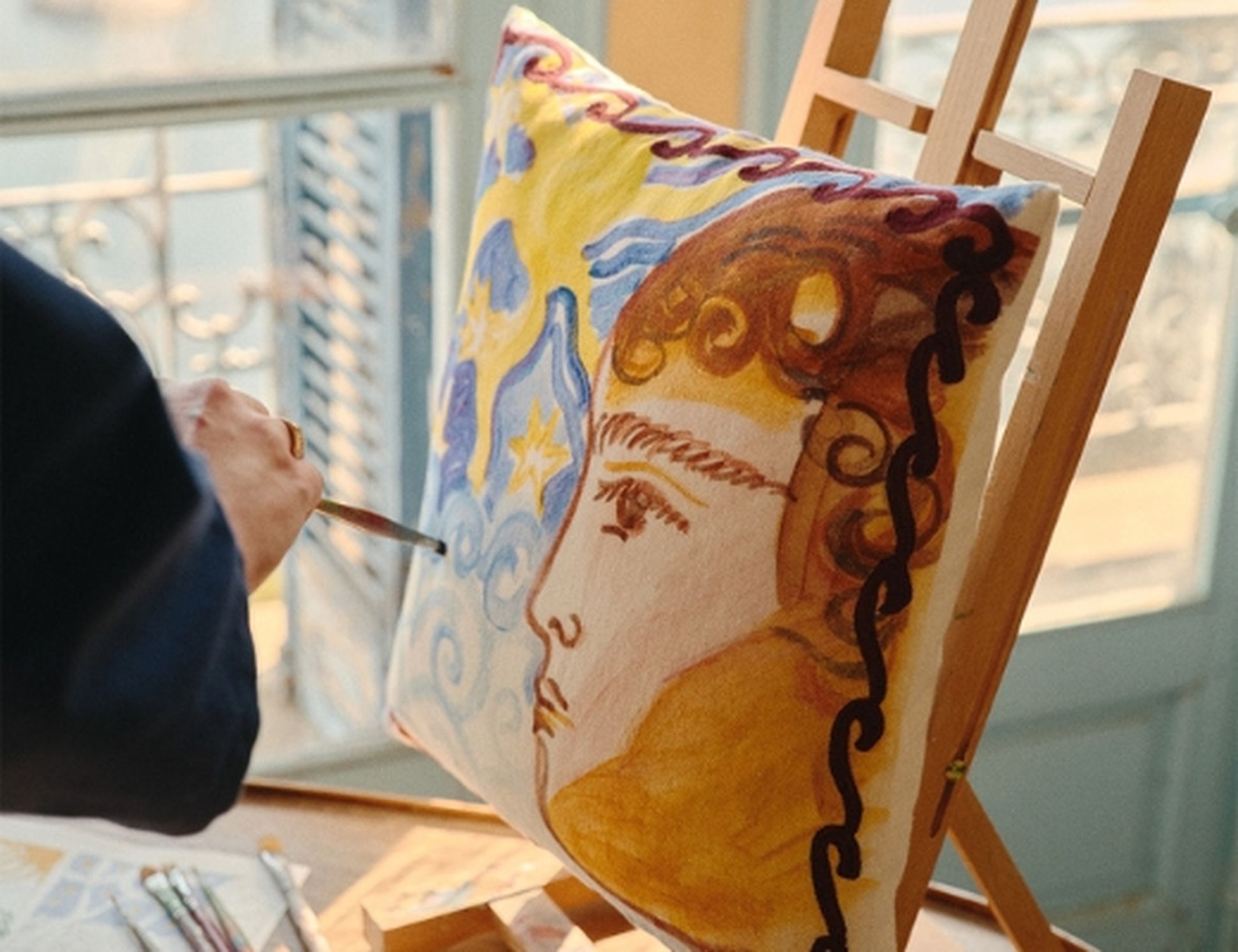 Peindre un portrait haut en couleur sur un coussin, posé sur un chevalet près d'une fenêtre ensoleillée avec vue sur la mer.