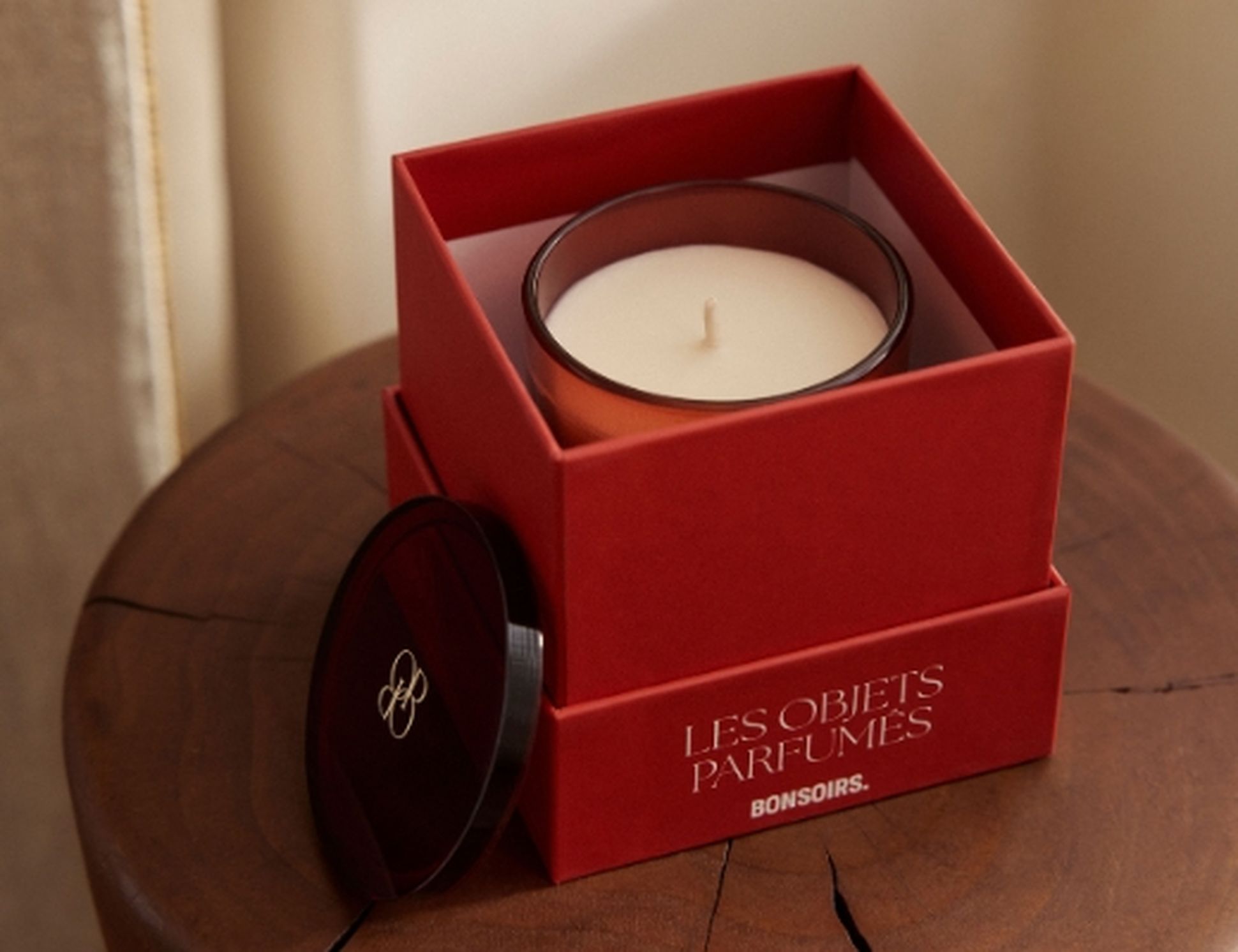 Boîte rouge contenant une bougie blanche, portant l'inscription « LES OBJETS PARFUMÉS, BONSOIRS », posée sur une surface en bois. Le couvercle repose à côté de la boîte.