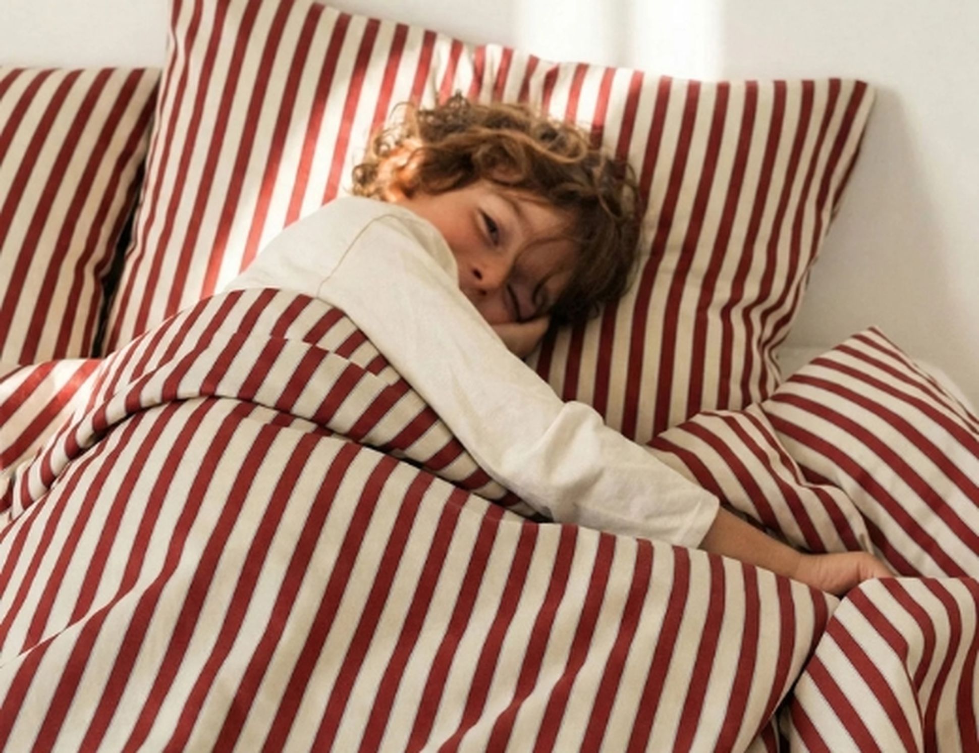 Un enfant allongé dans son lit, recouvert d'une parure de lit à rayures rouges et blanches, la tête posée sur des oreillers assortis, regardant vers l'appareil photo.