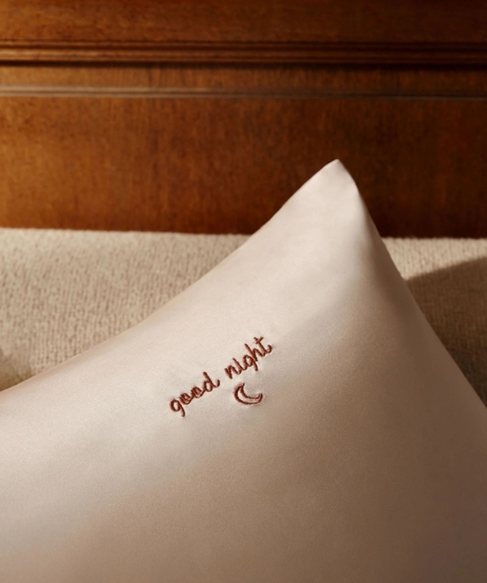 Un coussin crème portant la mention « good night » et un croissant de lune brodés en fil marron, posé sur un coussin beige près d'une tête de lit en bois.