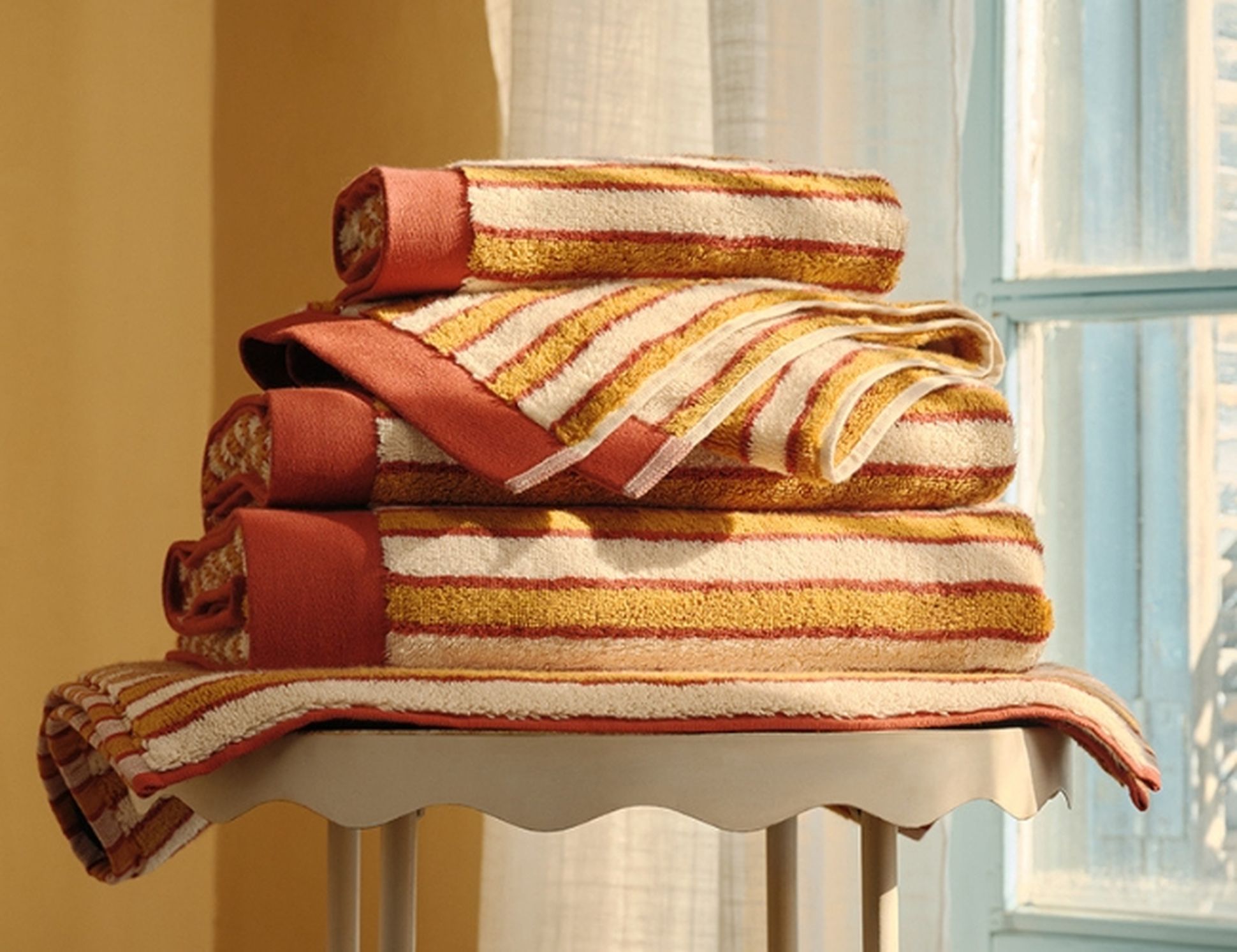 Une pile de serviettes rayées aux couleurs chaudes posée sur un tabouret près d'une fenêtre, baignée par la lumière du soleil, créant une ambiance chaleureuse.
