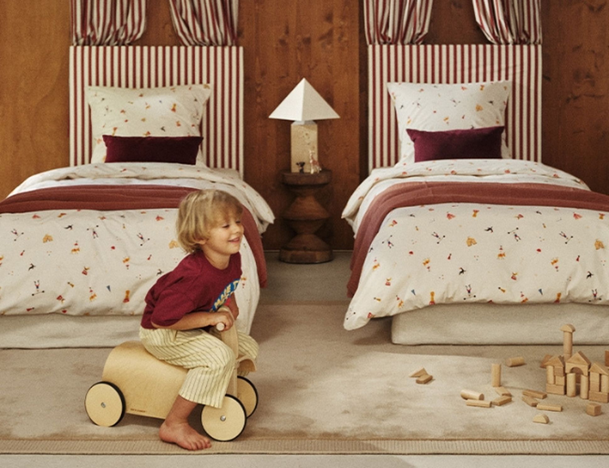 Un enfant monte un jouet en bois dans une chambre chaleureuse dotée de deux lits jumeaux, de ciels de lit à rayures rouges et blanches et d'un mur en bois en arrière-plan.