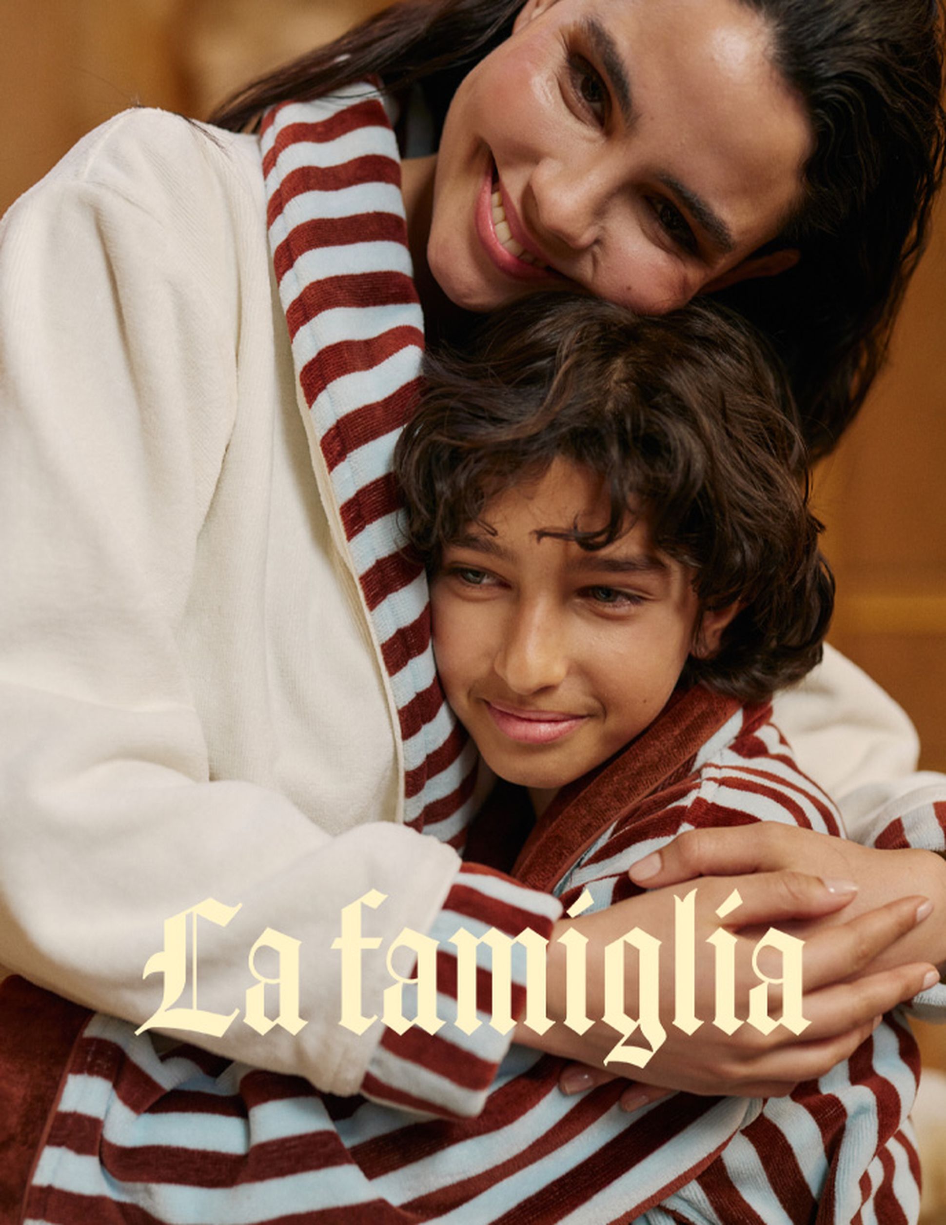 Une femme serre tendrement un enfant dans ses bras ; tous deux portent des vêtements rayés. Le texte « La famiglia » est superposé à l'image.