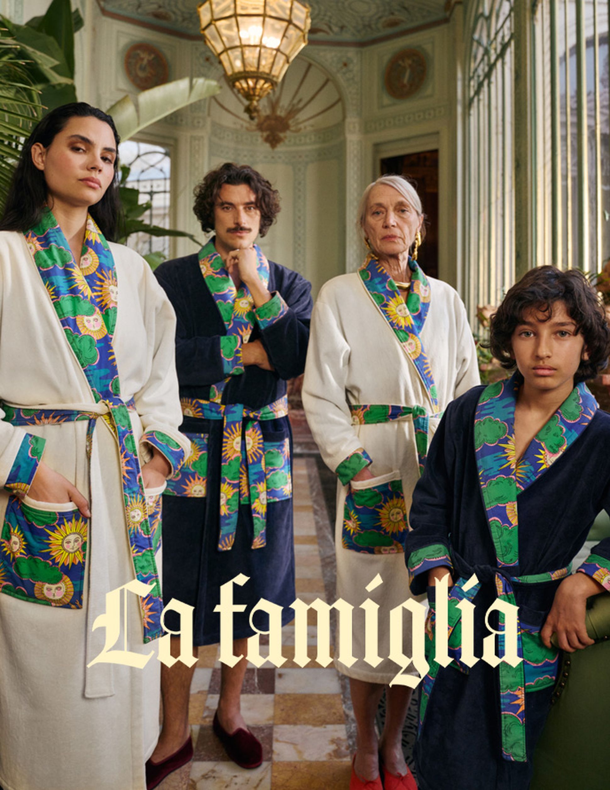 Quatre personnes vêtues de peignoirs colorés se tiennent fièrement dans une pièce richement décorée. Le texte indique « La famiglia ».