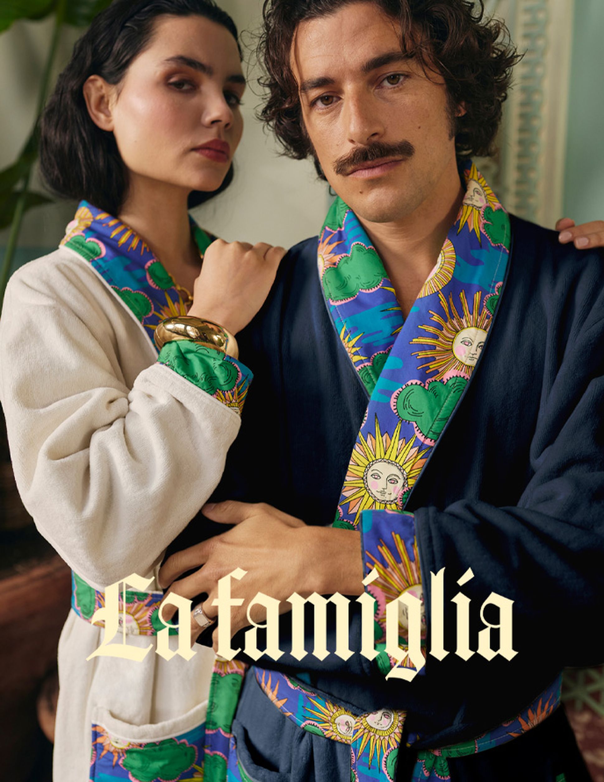 Un homme et une femme, vêtus de peignoirs aux motifs colorés, se tiennent côte à côte, le regard assuré. Le texte « La famiglia » apparaît en superposition sur l'image.