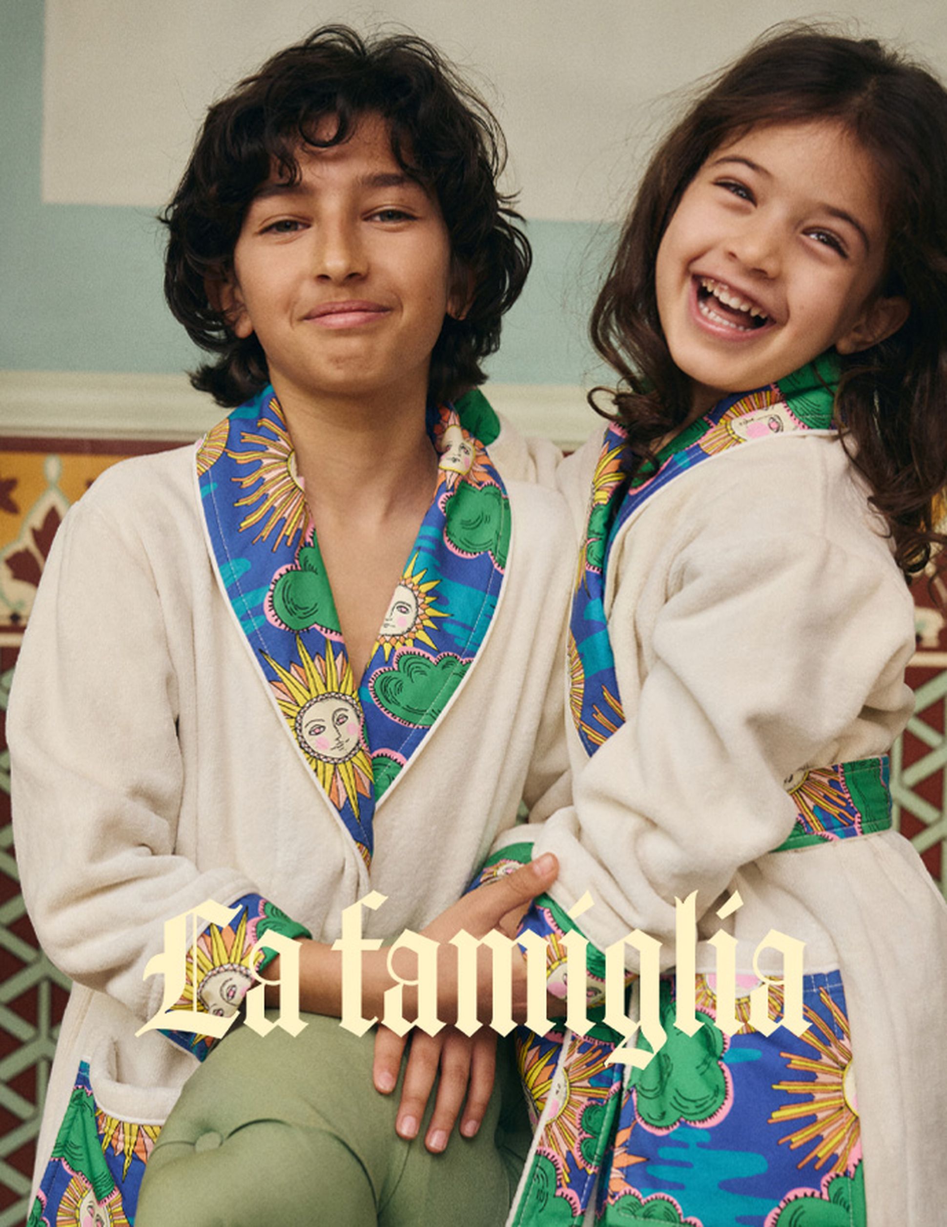 Deux enfants souriants, vêtus de peignoirs colorés ornés de motifs représentant des soleils et des nuages. Les mots « La famiglia » apparaissent en superposition sur l'image.