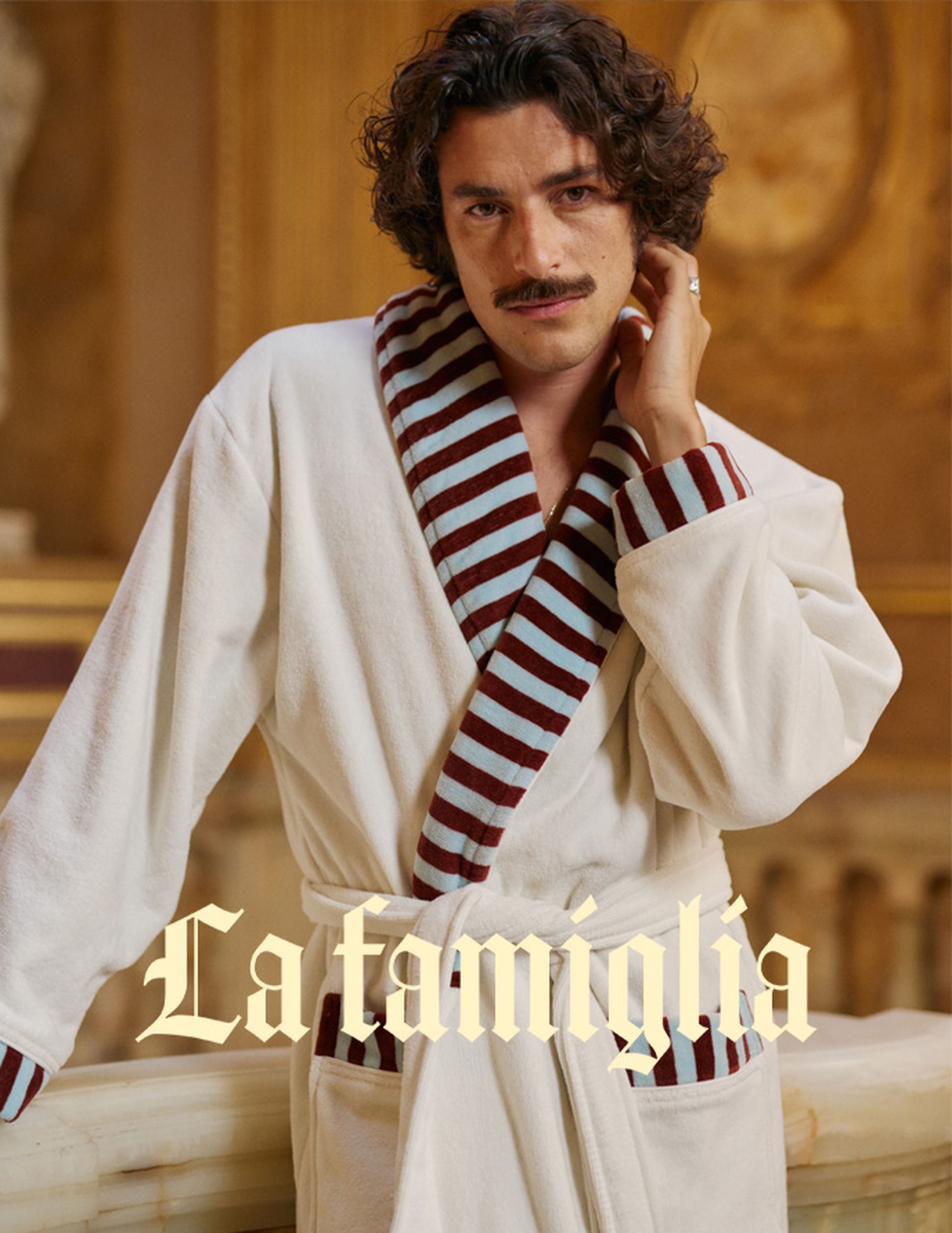 Un homme vêtu d'un peignoir blanc à rayures bordeaux se tient avec élégance à l'intérieur, avec le texte « La famiglia » superposé dans une police de caractères ornée.