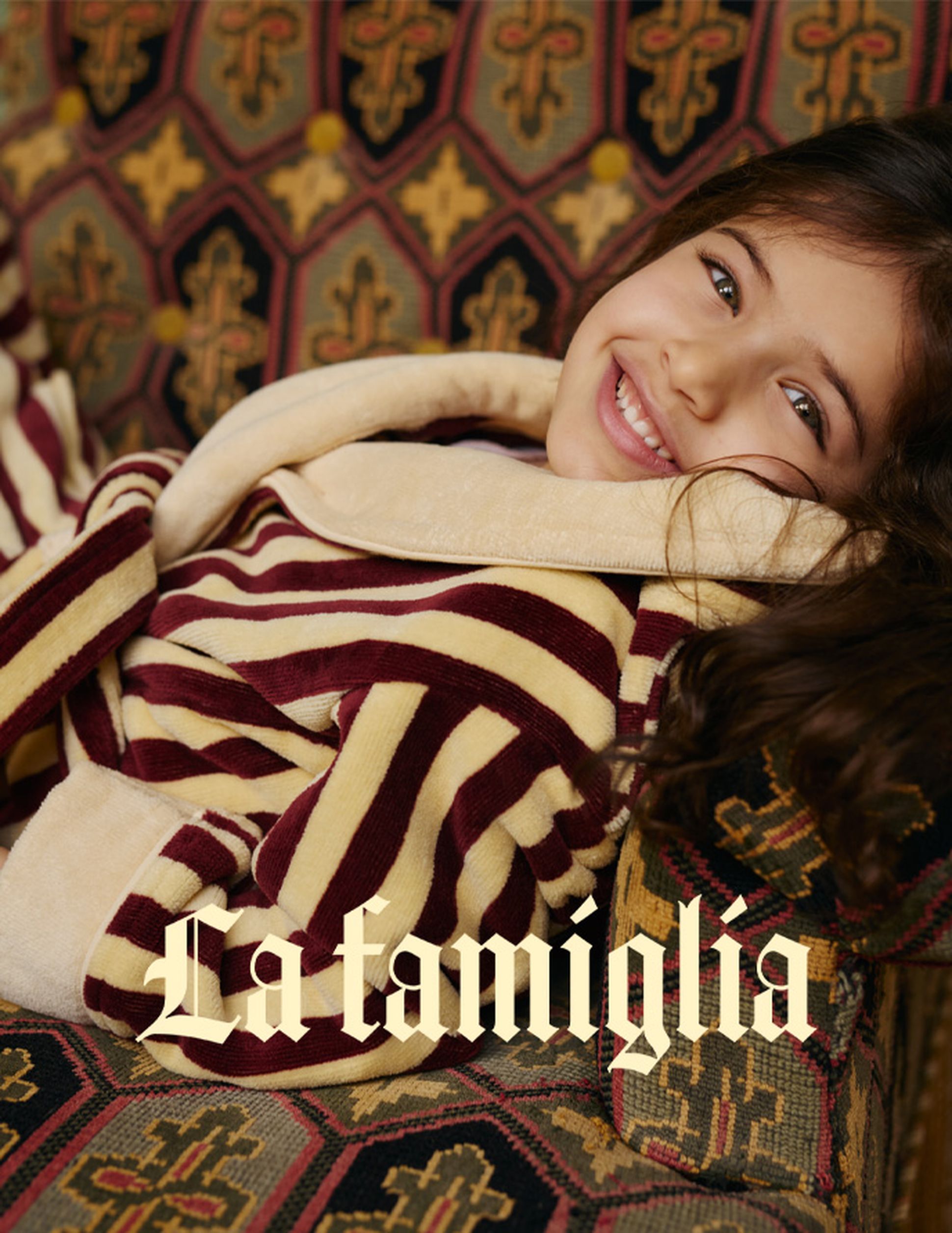 Un enfant souriant, vêtu d'un peignoir rayé, est allongé sur un coussin à motifs. Le texte « La famiglia » apparaît en superposition sur l'image.
