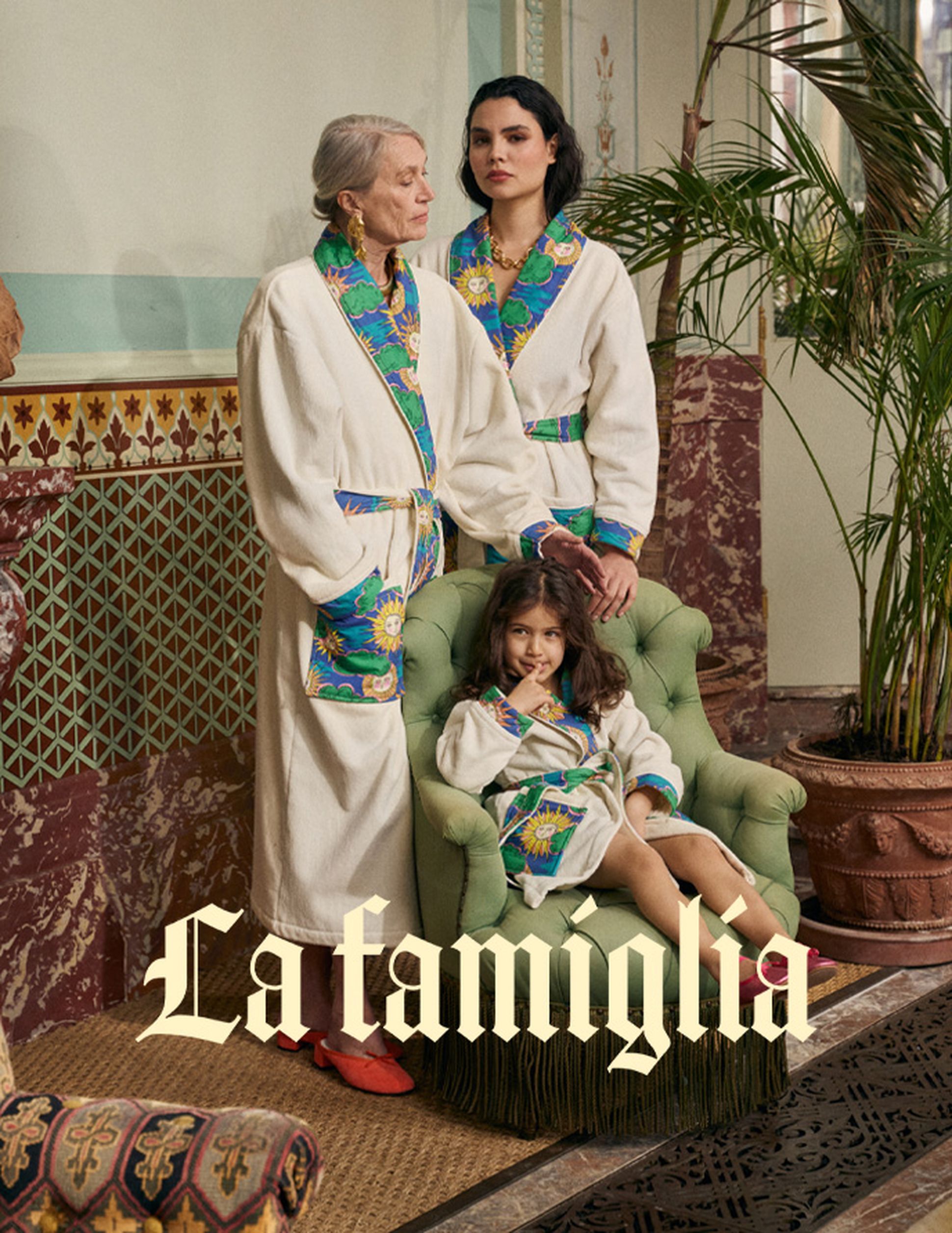 Trois personnes vêtues de peignoirs assortis aux motifs colorés sont debout ou assises dans une pièce élégante, agrémentée de plantes et de murs à motifs. On peut lire « La famiglia »