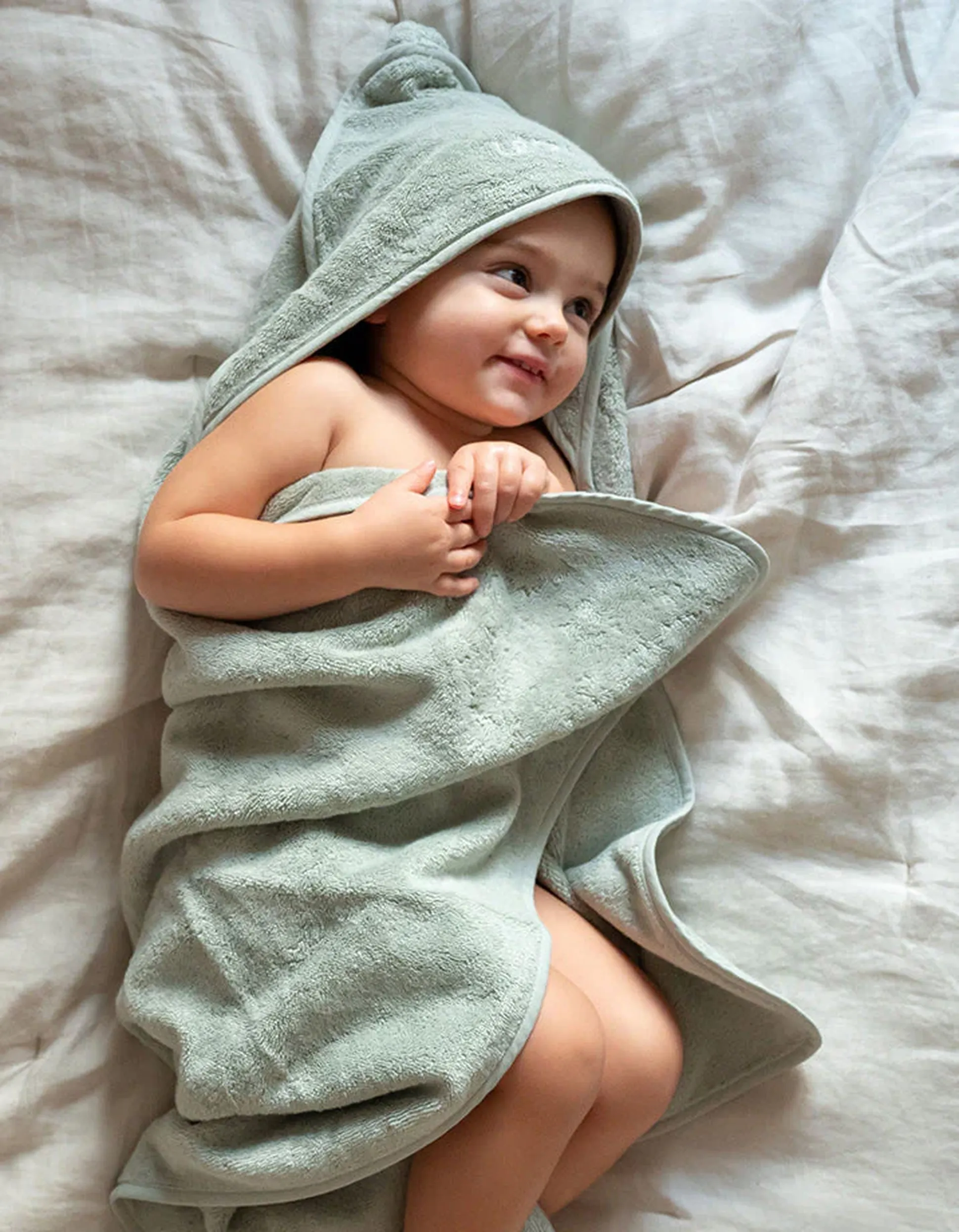 Un bébé est enveloppé dans une serviette à capuche douce et vert clair ; il est allongé sur un lit et semble content.