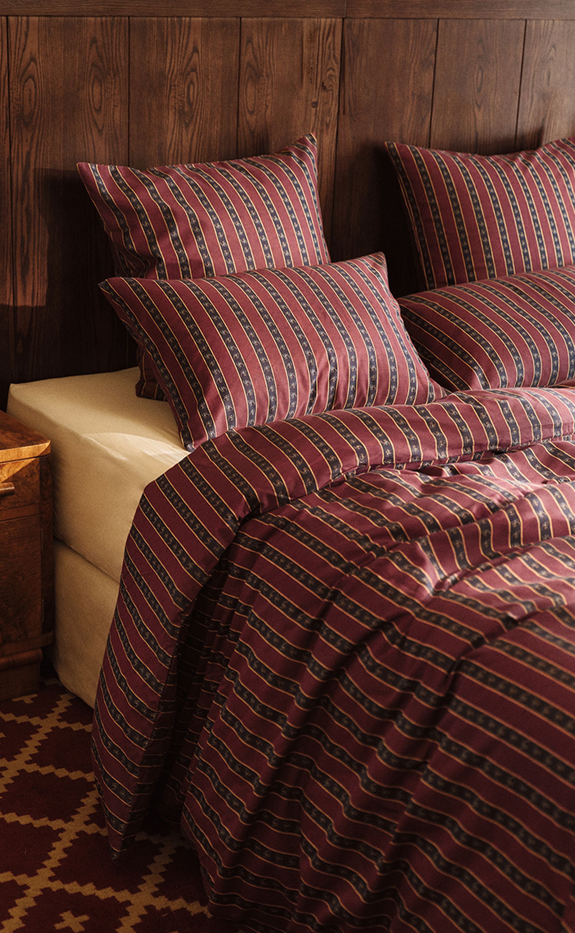 Un lit recouvert d'une parure de lit à rayures rouge foncé et bleu marine, comprenant des oreillers et une couette, adossé à un mur en bois, à côté d'une table de chevet en bois.