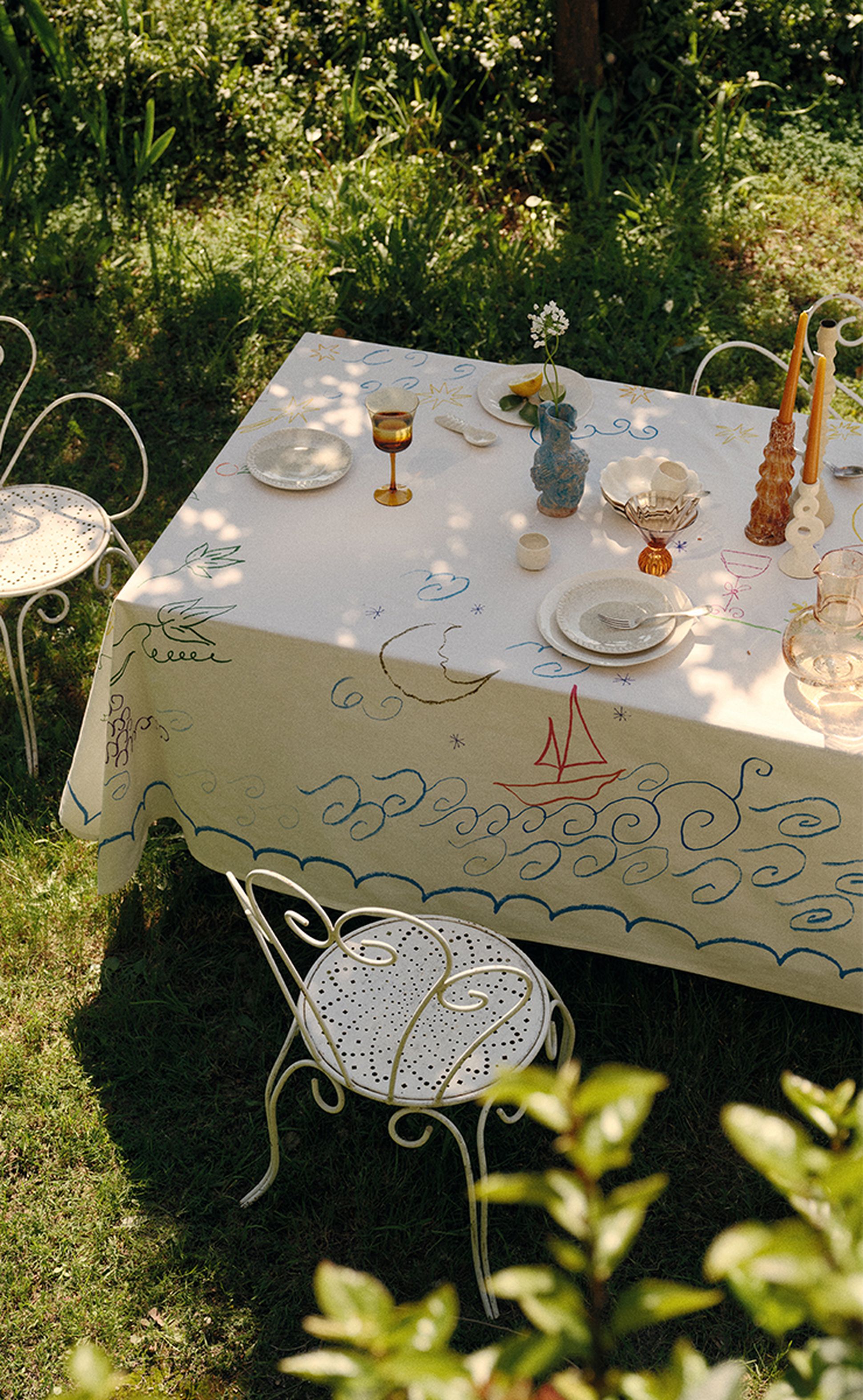 Une table d'extérieur recouverte d'une nappe sur le thème de la mer, dressée avec des verres et des couverts, entourée de chaises en fer forgé blanc sur une pelouse verte.