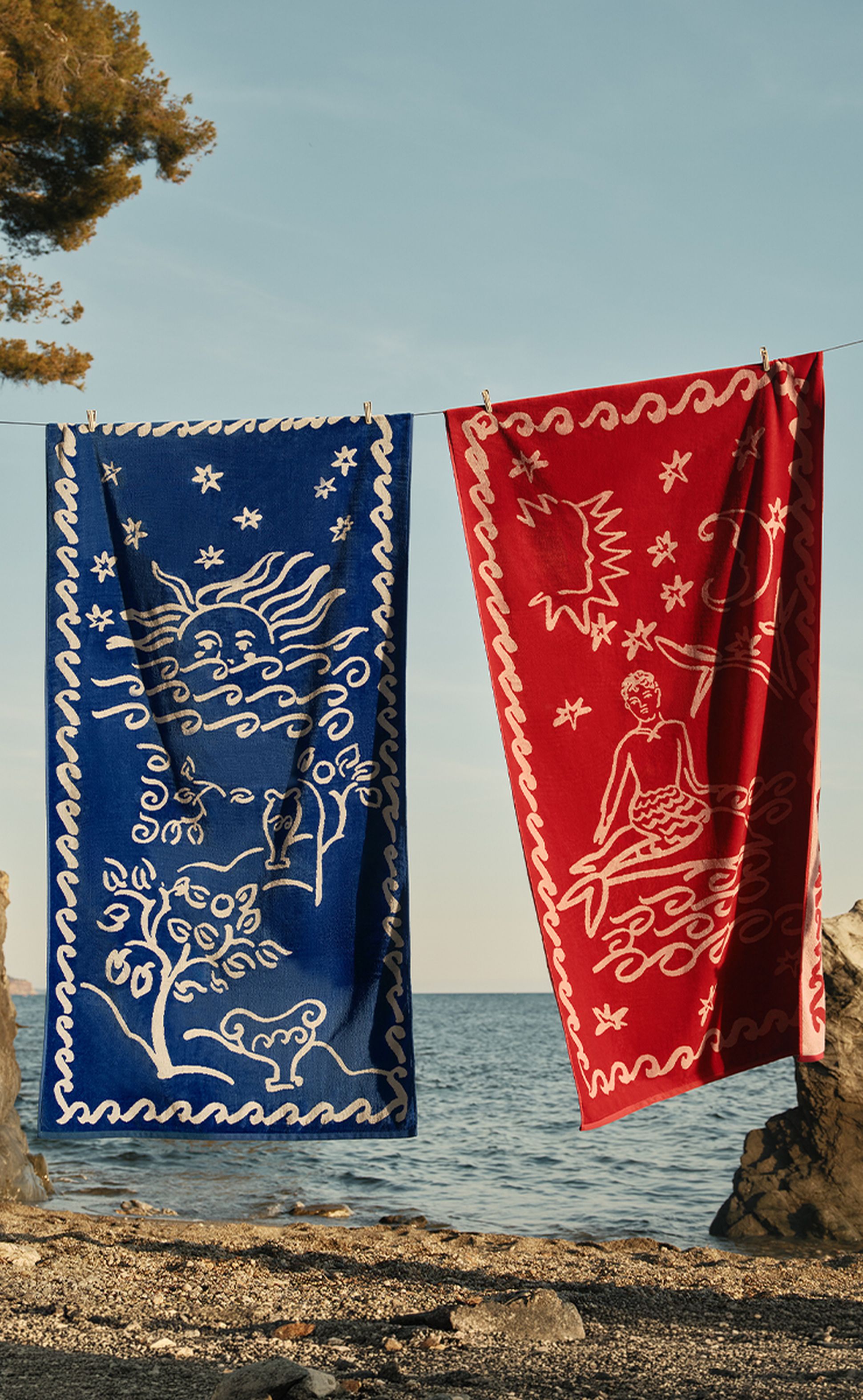 Deux serviettes aux couleurs vives, l'une bleue et l'autre rouge, ornées de motifs blancs inspirés du ciel et de l'océan, sont suspendues à une corde près de la mer.