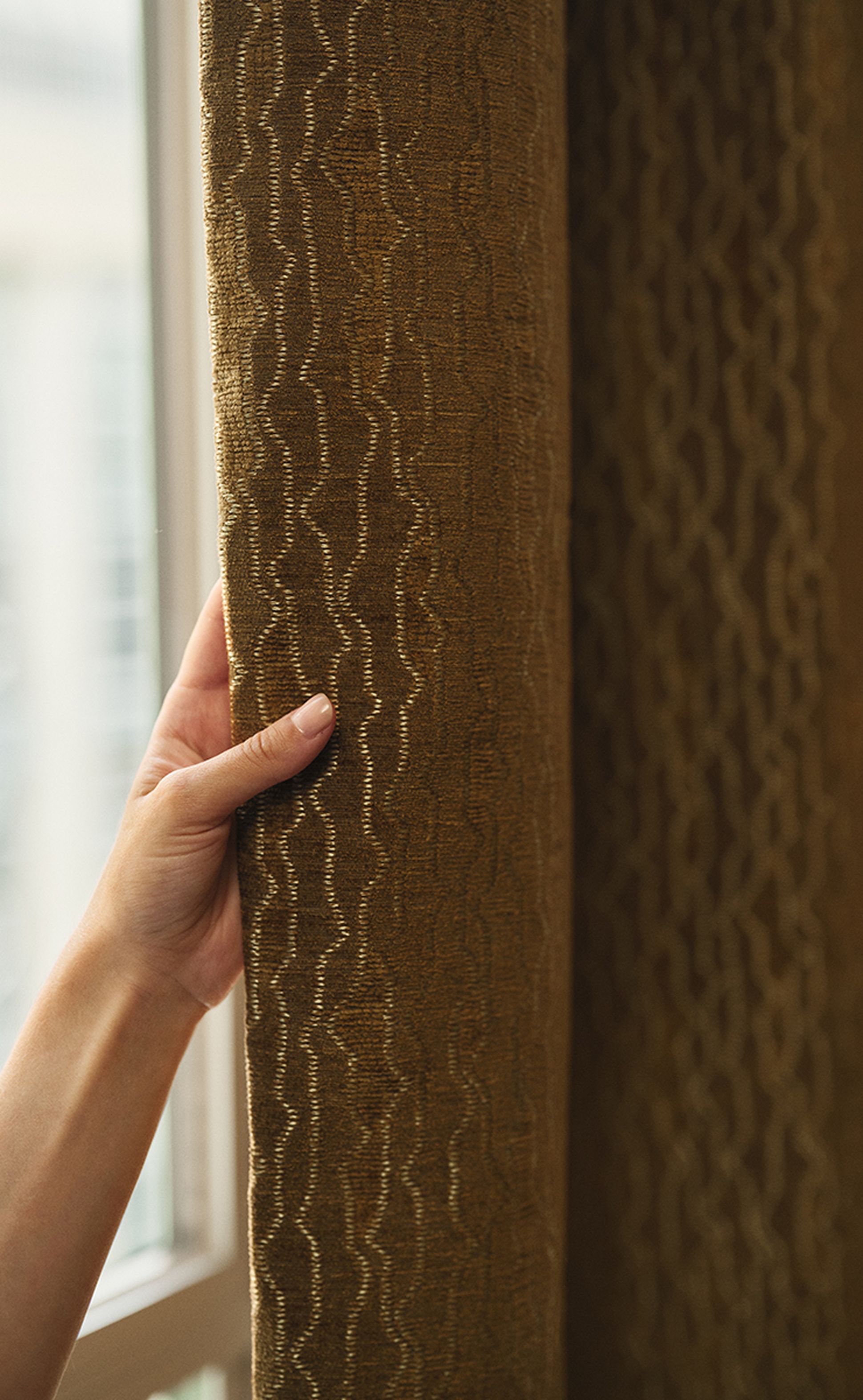 Une main tenant un rideau marron texturé aux motifs ondulés, près d'une fenêtre.