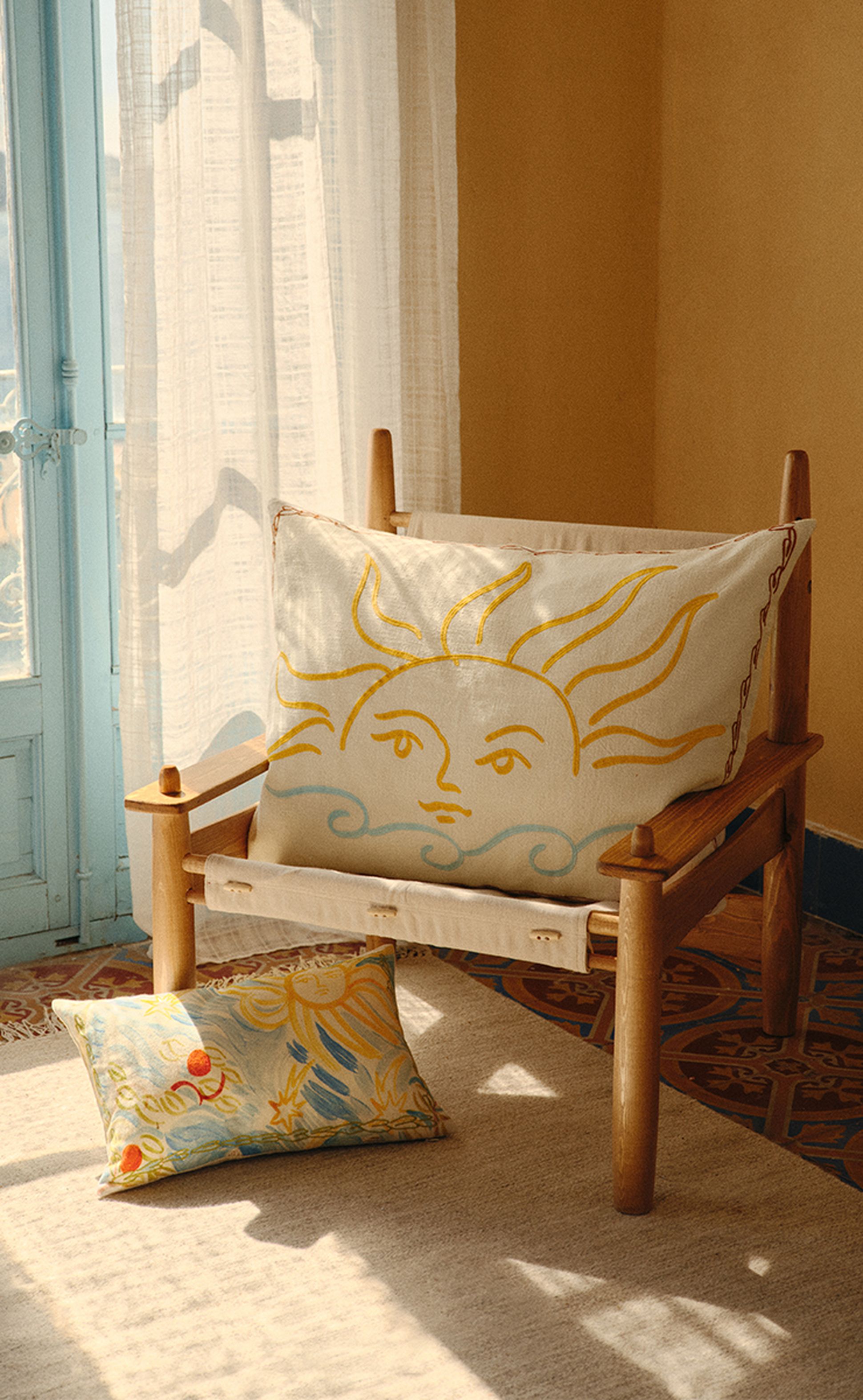 Chaise en bois avec un coussin sur le thème du soleil, baignée d'une lumière chaude près d'une fenêtre à encadrement bleu, posée sur un sol en carrelage à motifs.