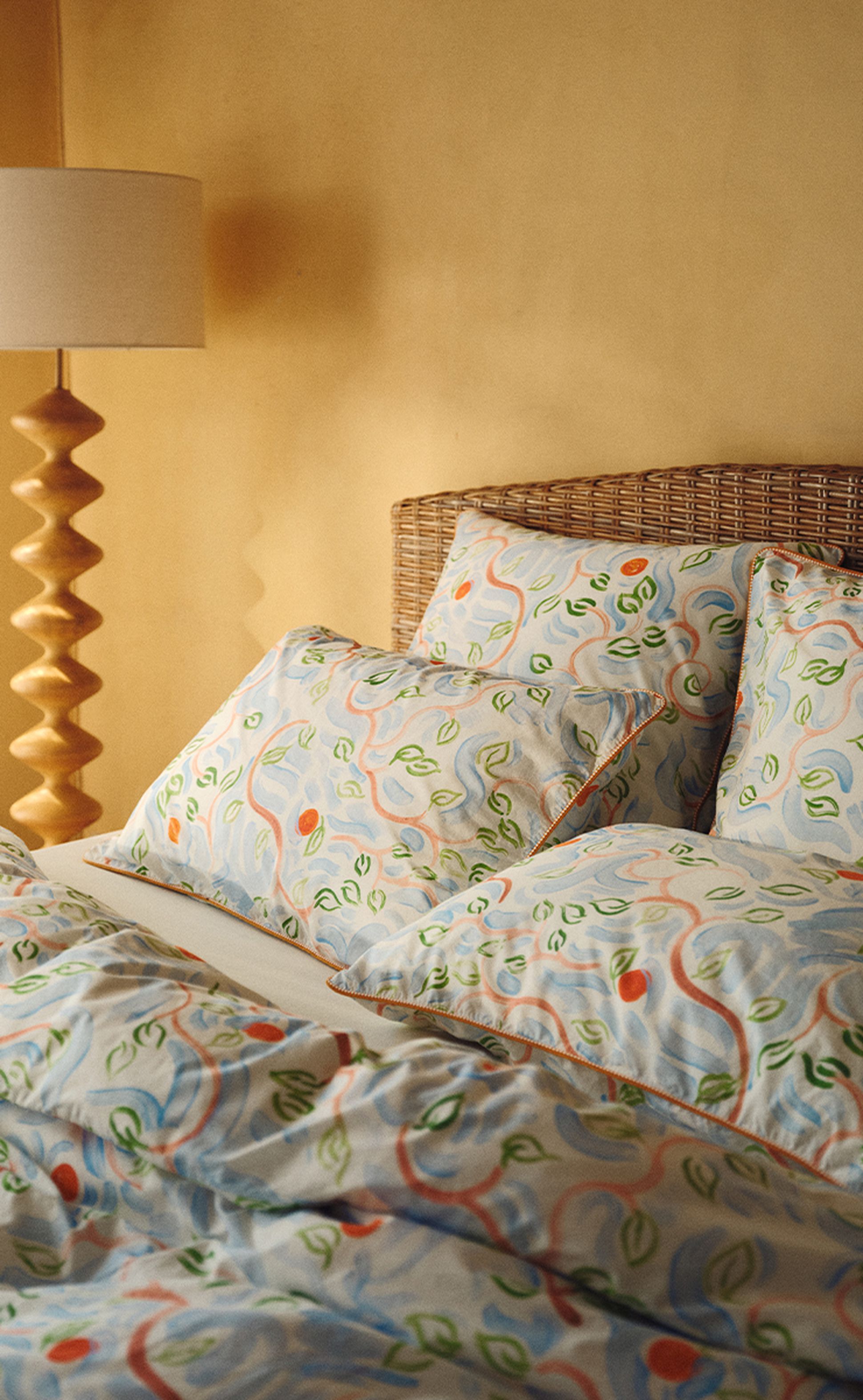 Un lit avec des draps et des oreillers aux motifs de feuilles colorés, une tête de lit en osier et une lampe de chevet en bois, devant un mur beige aux tons chauds.