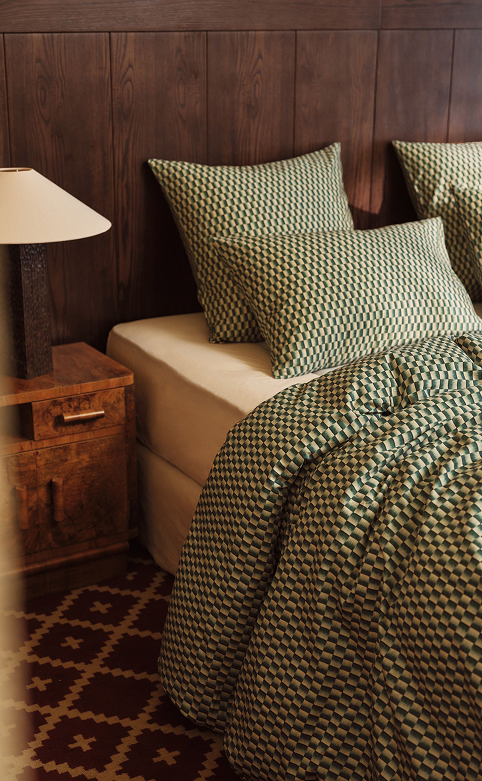 Cette chambre confortable comprend un lit avec une parure de lit à motifs verts et blancs, des oreillers assortis, une tête de lit en bois, une table de chevet et une lampe.