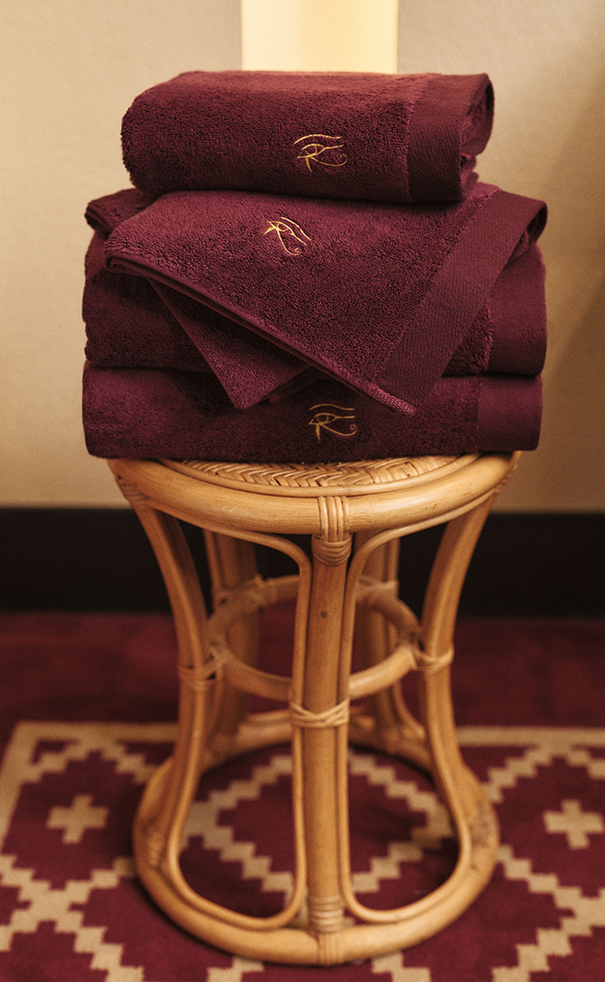 Une pile de serviettes bordeaux ornées de broderies dorées, posée sur un tabouret en osier, au milieu d'un tapis à motifs.