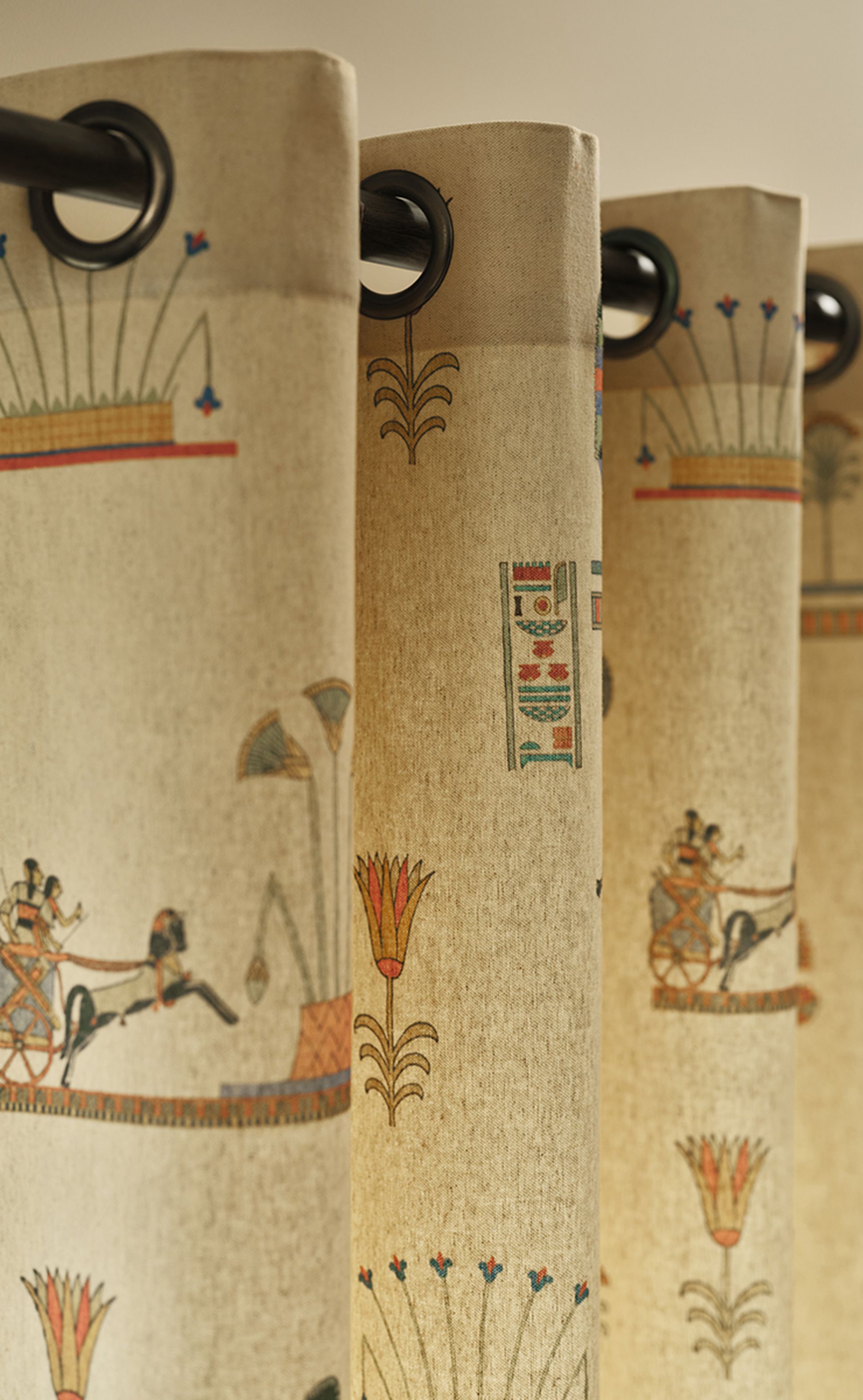Des rideaux beiges ornés de motifs de style égyptien en rouge, bleu et vert, représentant des chars et des fleurs de lotus, suspendus à une tringle noire.