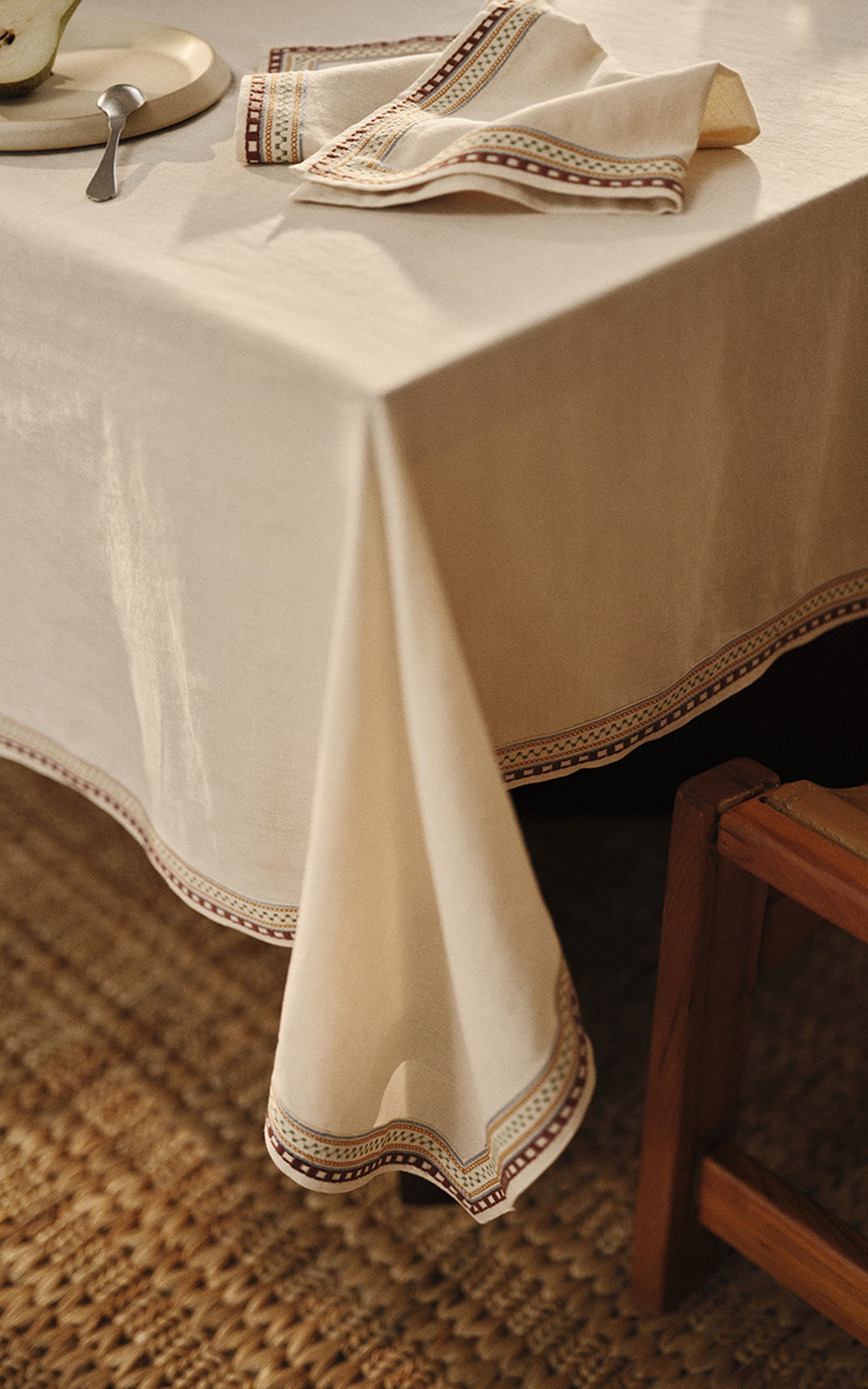 Une table à manger dressée avec une nappe crème, des assiettes, des bougies, des verres et des bols en bois, entourée de chaises en bois, dans une pièce baignée d'une lumière tamisée.