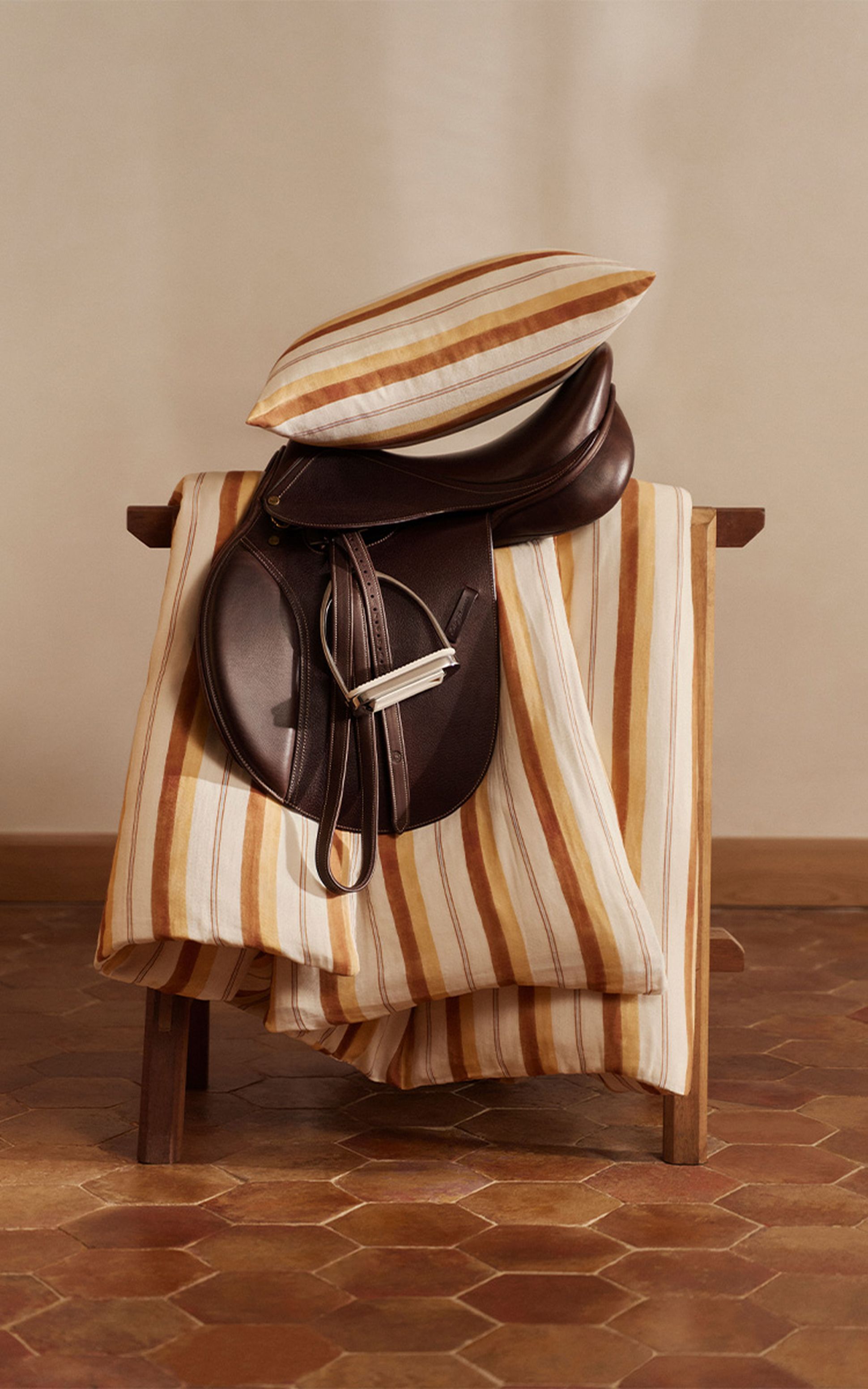 Une selle en cuir poli et des textiles rayés sont élégamment disposés sur un banc en bois, sur fond de sol carrelé.