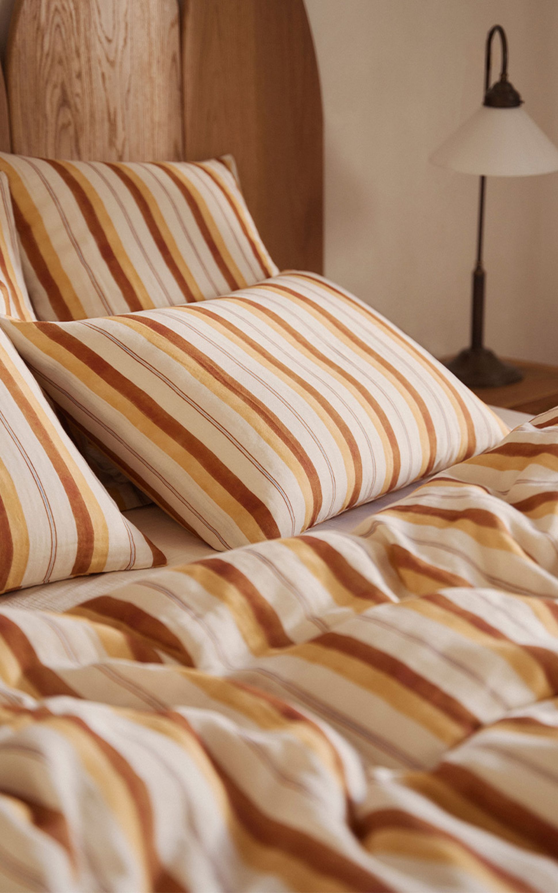 Un lit douillet avec des oreillers rayés et une couette dans les tons marron, orange et blanc. Une tête de lit en bois et une lampe se trouvent à l'arrière-plan.