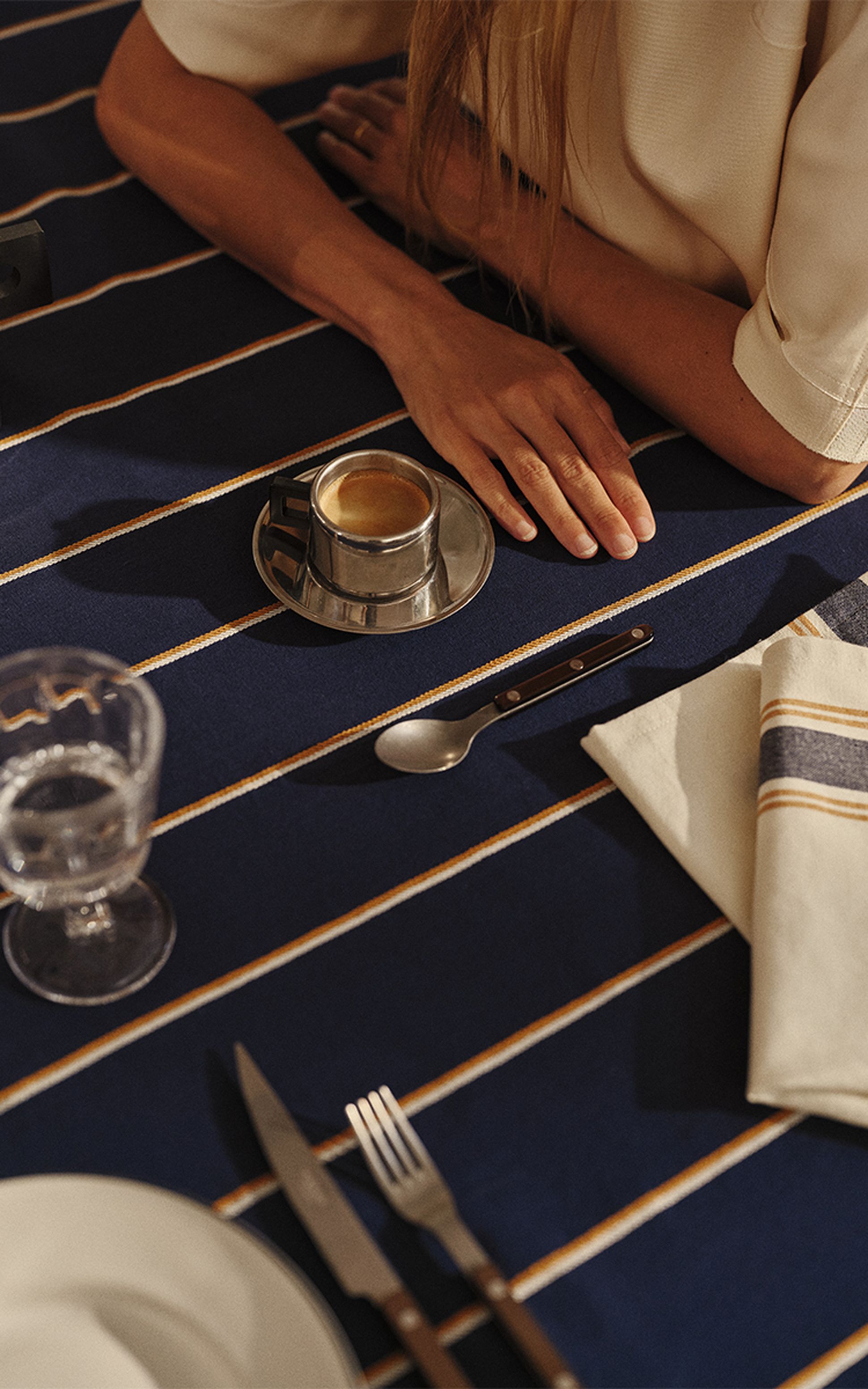 Une personne est assise à une table bleue rayée, devant laquelle sont soigneusement disposés une tasse d'expresso, des couverts, une serviette et un verre d'eau.