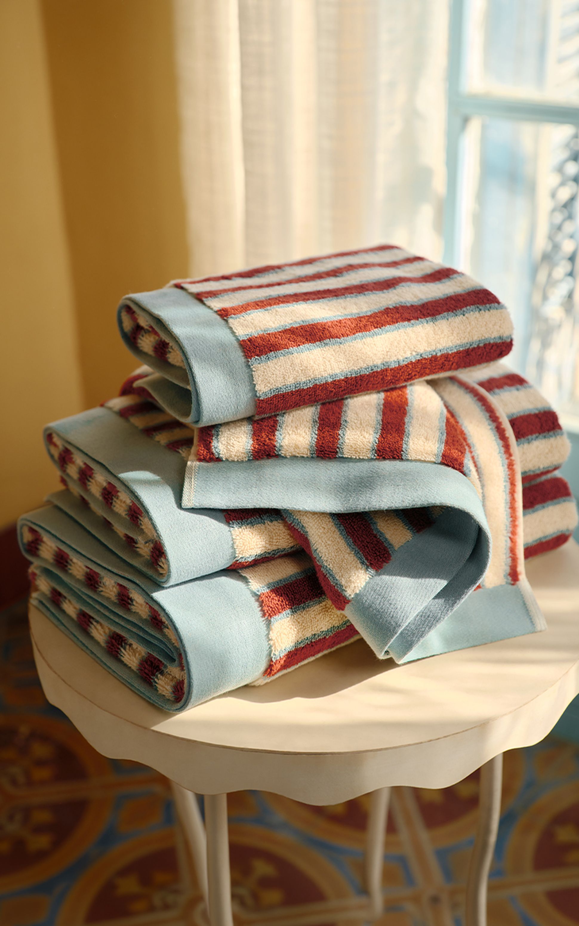 Une pile de serviettes rayées rouges, bleues et beiges posée sur une table ronde blanche, dans une pièce baignée de soleil au sol à motifs.