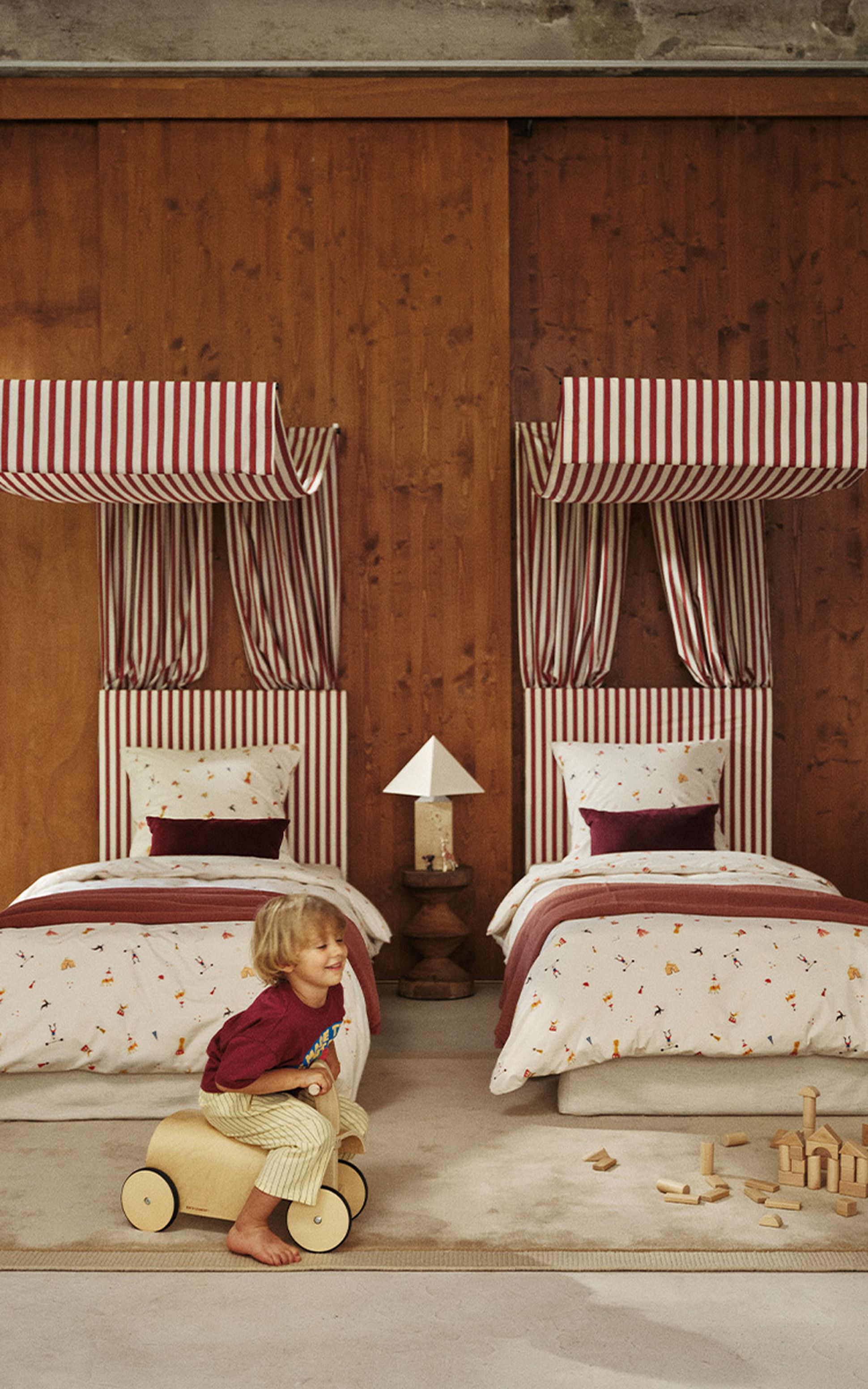 Un enfant joue avec un petit véhicule dans une chambre douillette équipée de deux lits, ornés de baldaquins rayés et d'une literie aux motifs ludiques, devant un mur lambrissé.