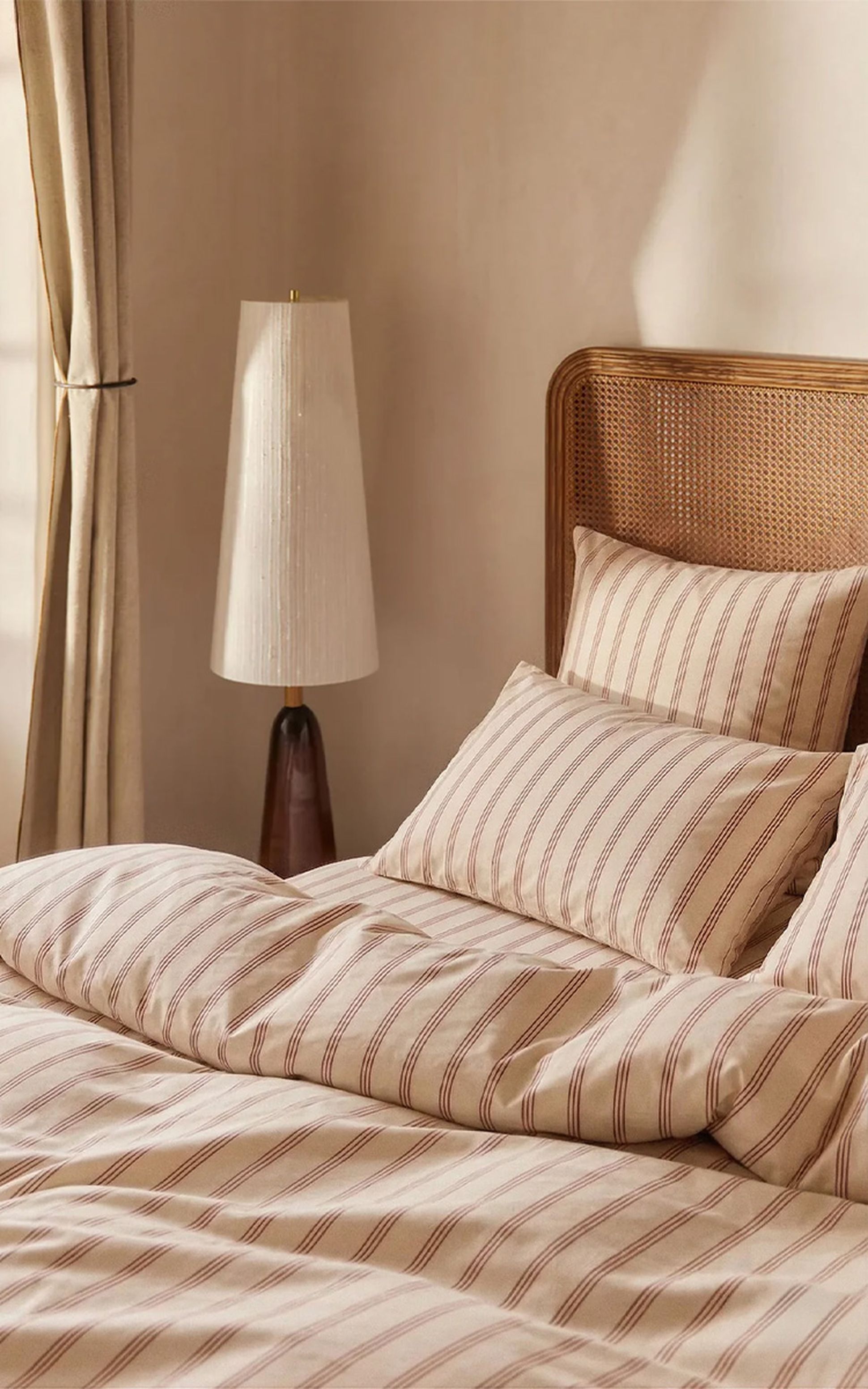 Une chambre douillette avec un lit à tête de lit en osier, une literie rayée toute douce et un lampadaire en bois à côté, le tout baigné d'une douce lumière naturelle.
