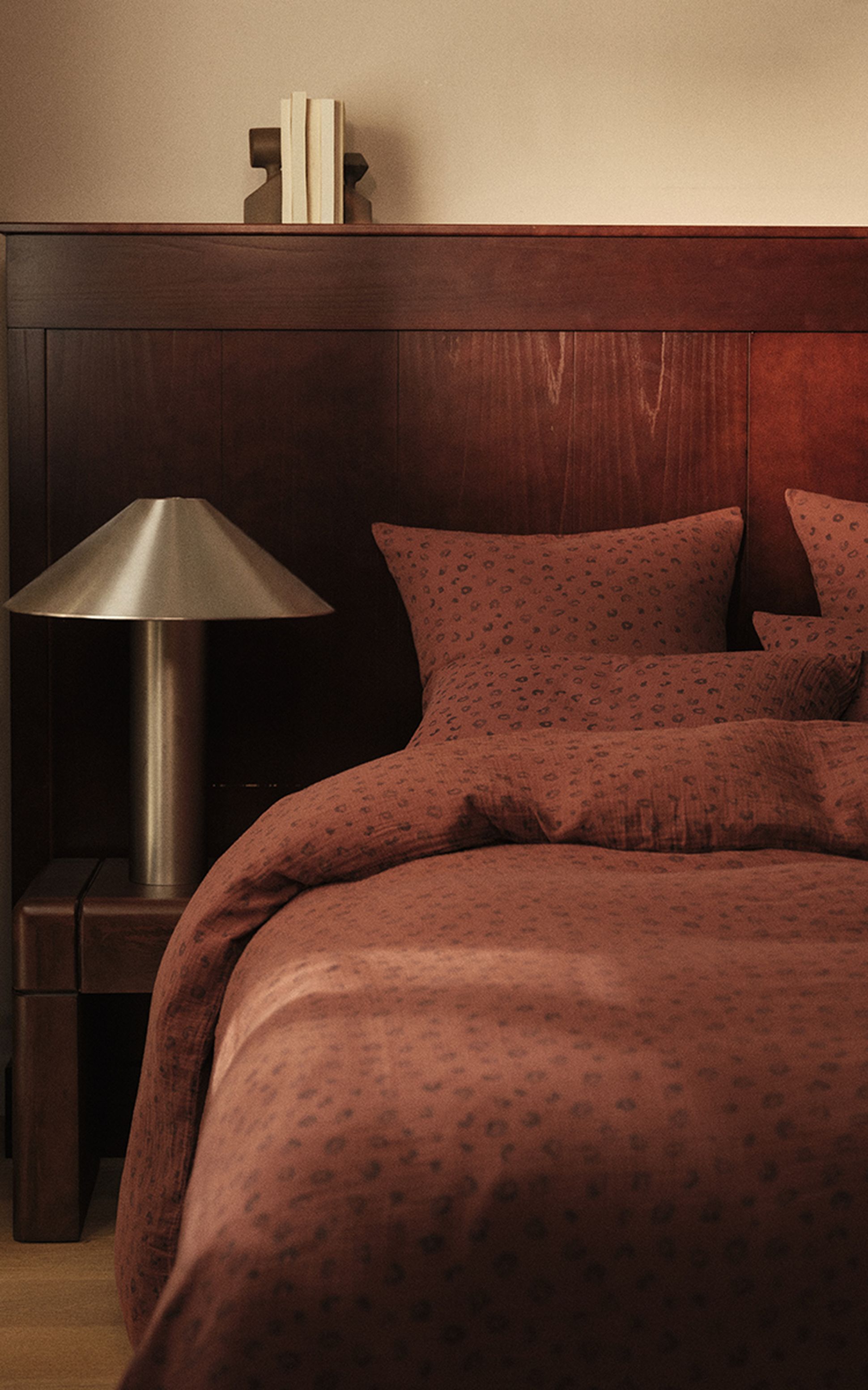 Une chambre douillette avec une tête de lit en bois, une parure de lit à motifs marron, une table de chevet, une lampe moderne et quelques livres sur une étagère.