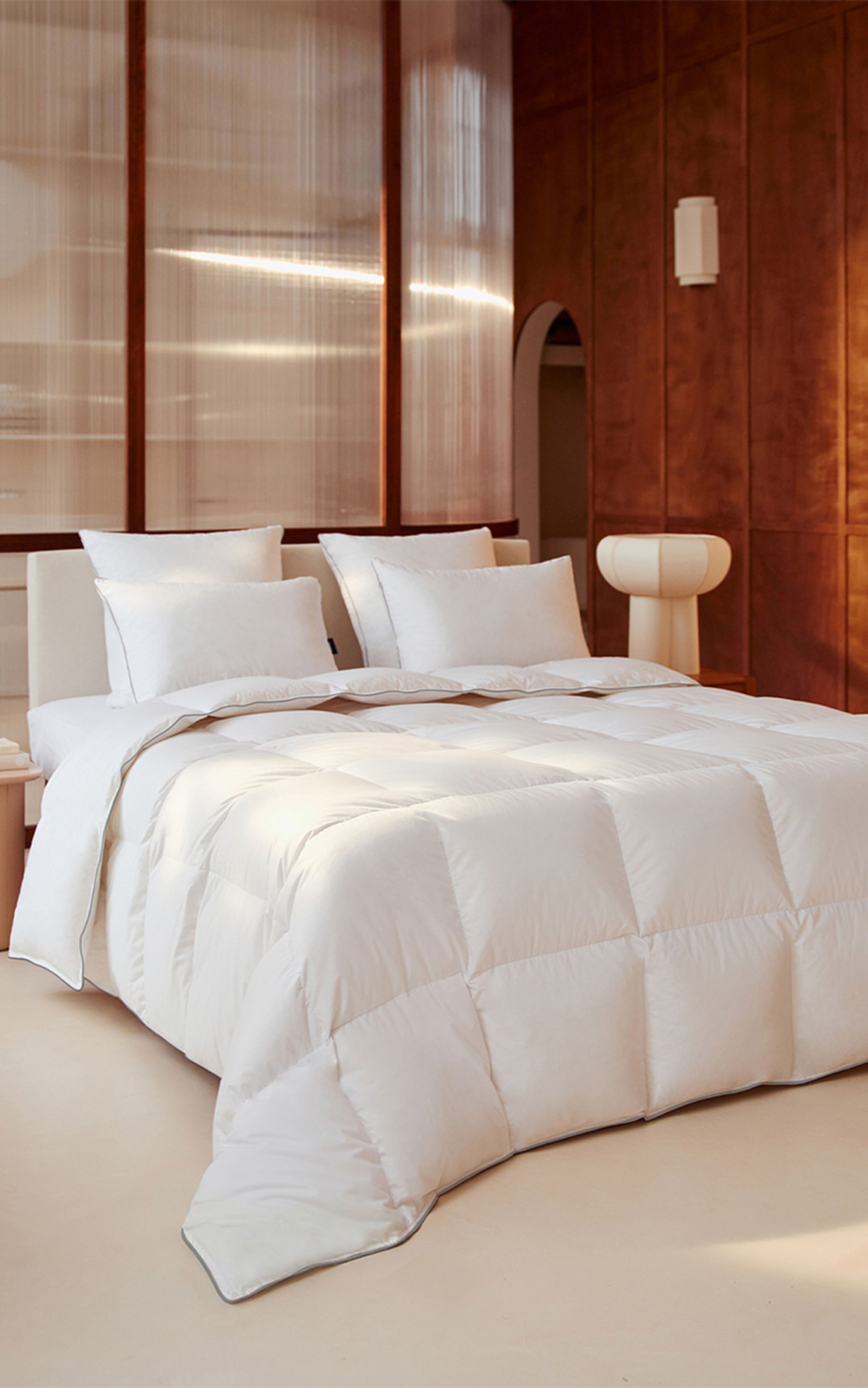 Un lit blanc douillet, garni d'oreillers moelleux et d'une couette bien gonflée, dans une chambre moderne à l'éclairage chaleureux, agrémentée de touches de bois et d'une chaise minimaliste.