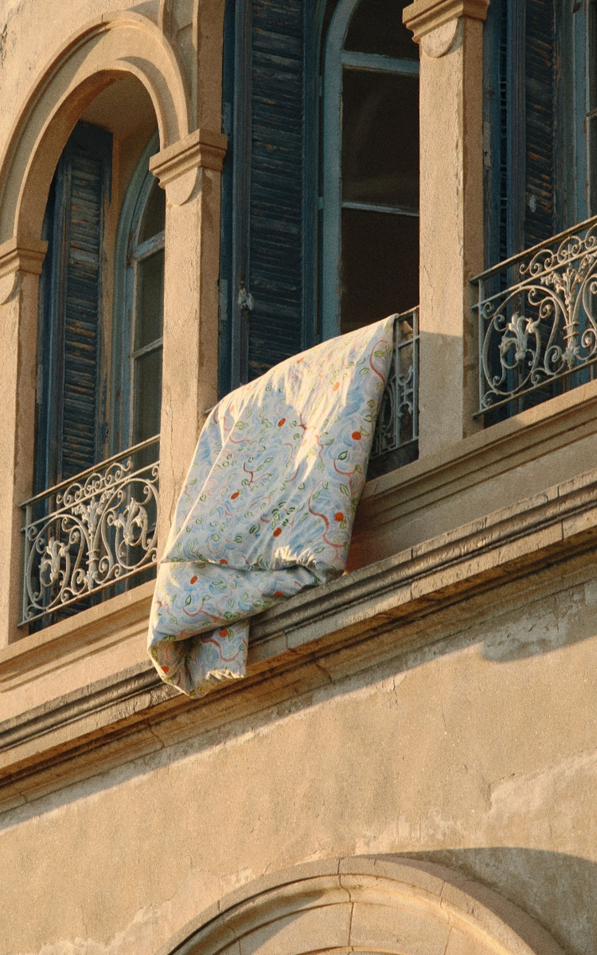 Une courtepointe colorée est suspendue à la balustrade en fer forgé d'un balcon, sur un immeuble aux fenêtres cintrées et aux volets bleus.