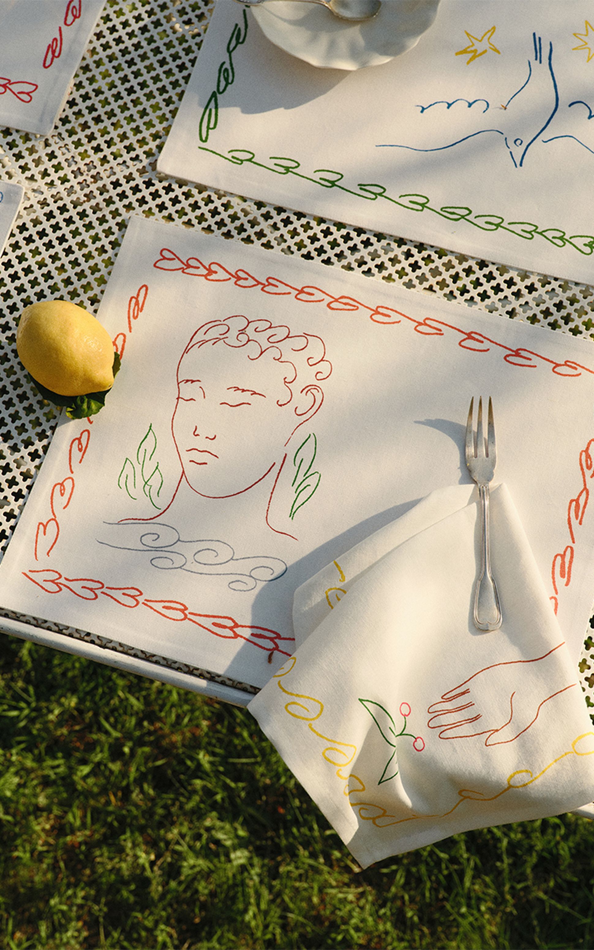 Une nappe décorative ornée de visages dessinés au trait, d'un citron et d'une fourchette posée sur une serviette, le tout disposé sur une table d'extérieur à motifs.