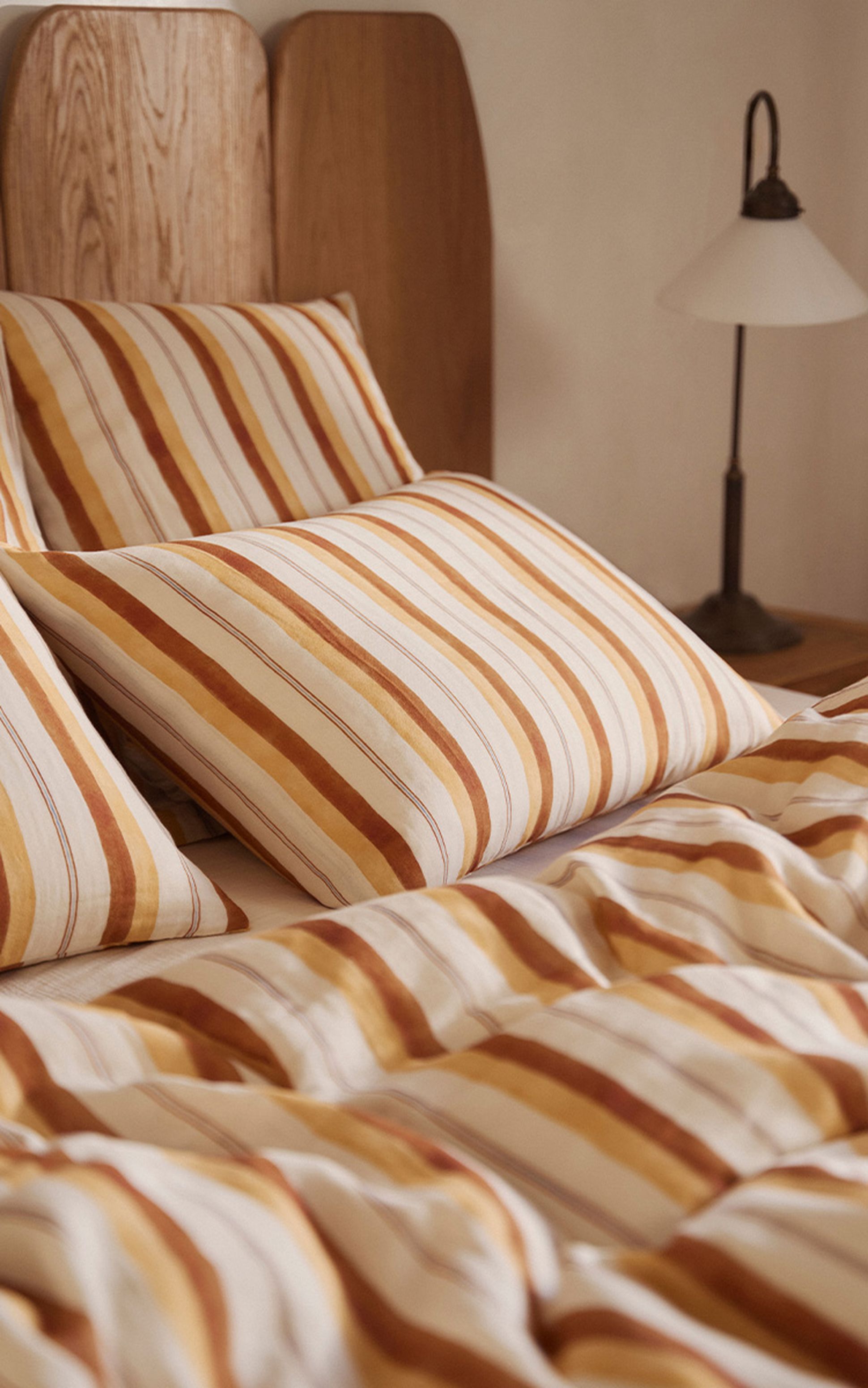 Oreillers rayés et literie aux tons chauds sur un lit en bois, avec une lampe de chevet en arrière-plan.