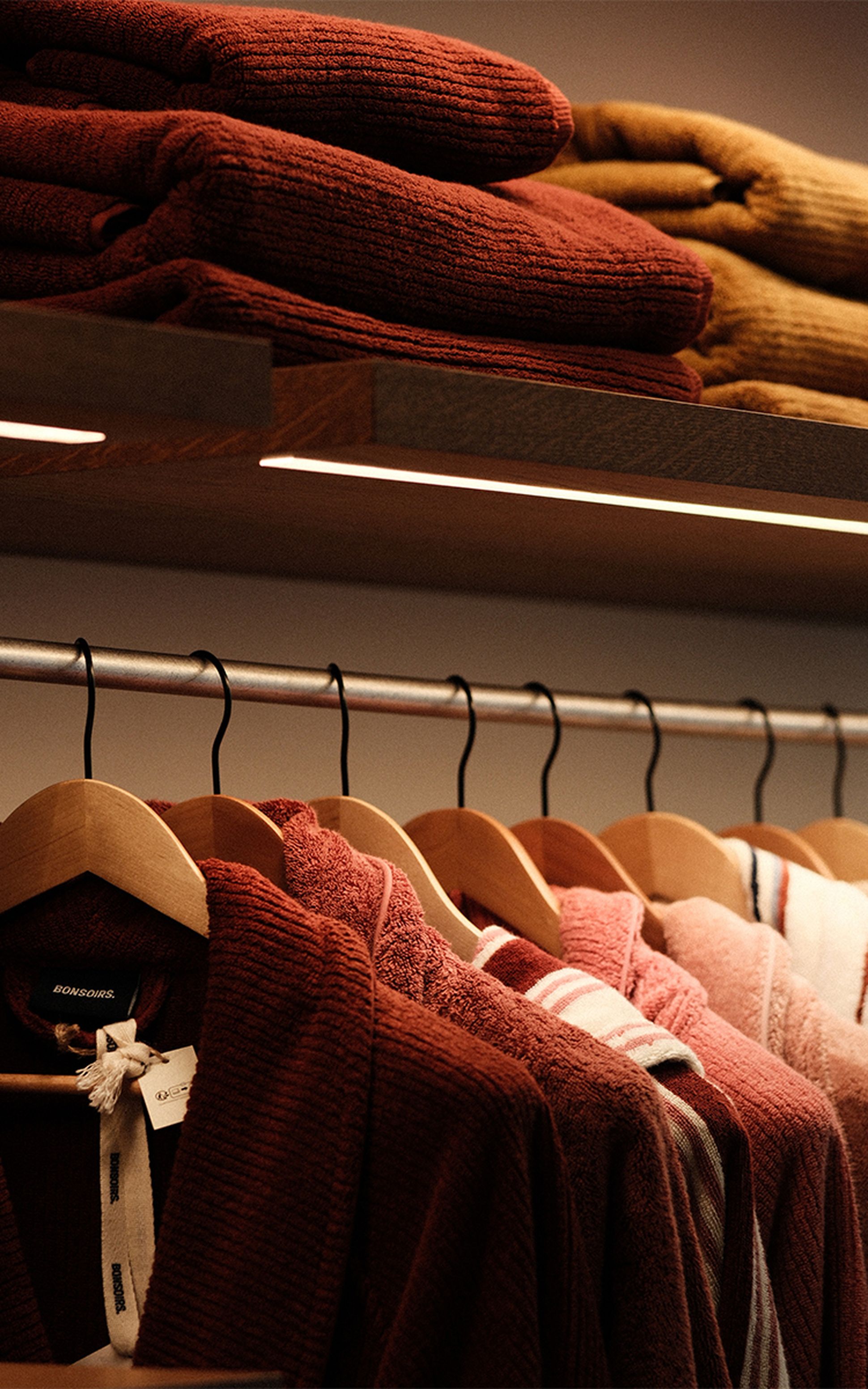 Une rangée de peignoirs rouges et roses suspendus à des cintres en bois, avec des serviettes pliées empilées sur une étagère au-dessus.