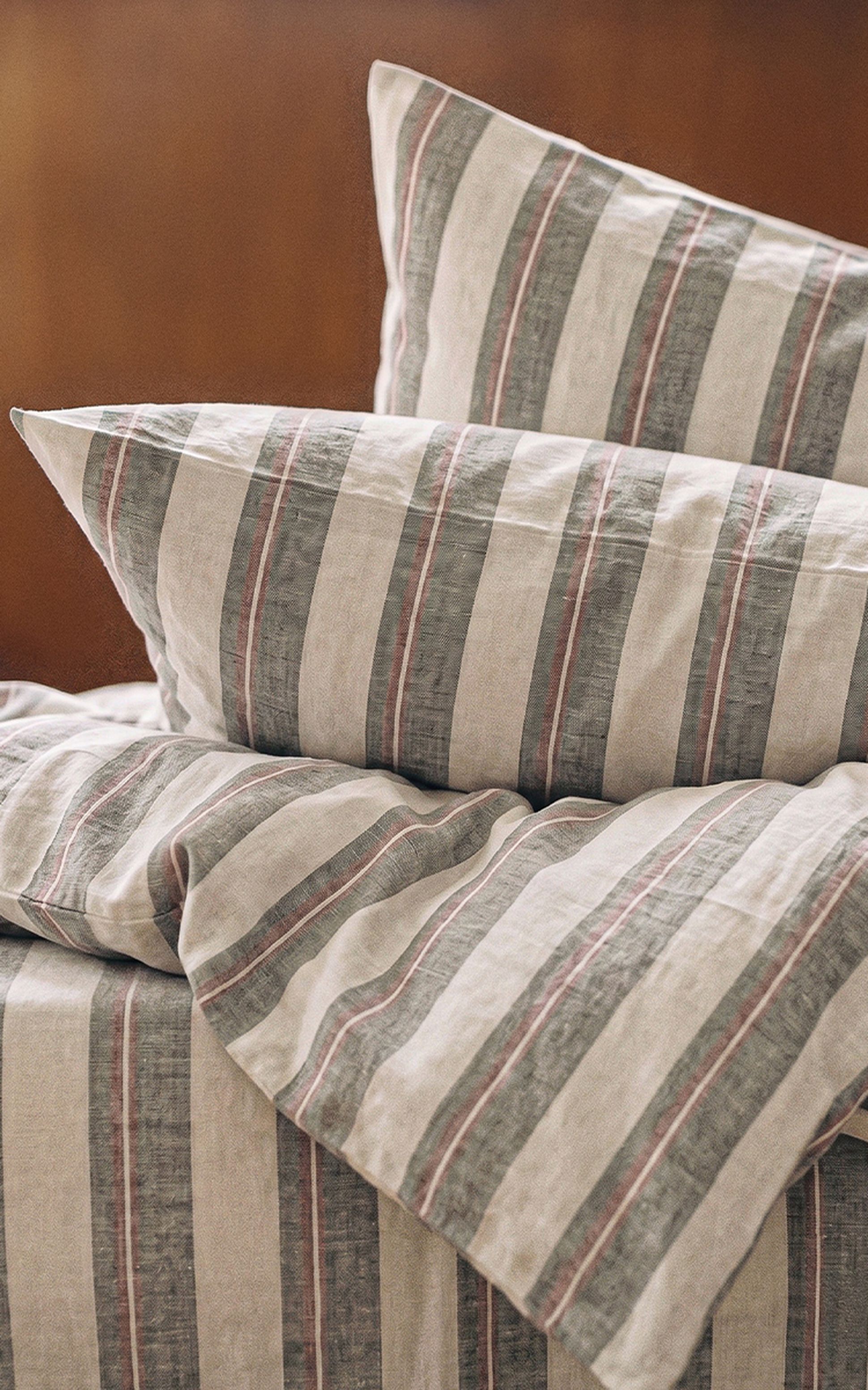 Gros plan sur deux oreillers et une couverture à rayures verticales beiges, grises et rouges, posés sur un lit doté d'une tête de lit en bois.