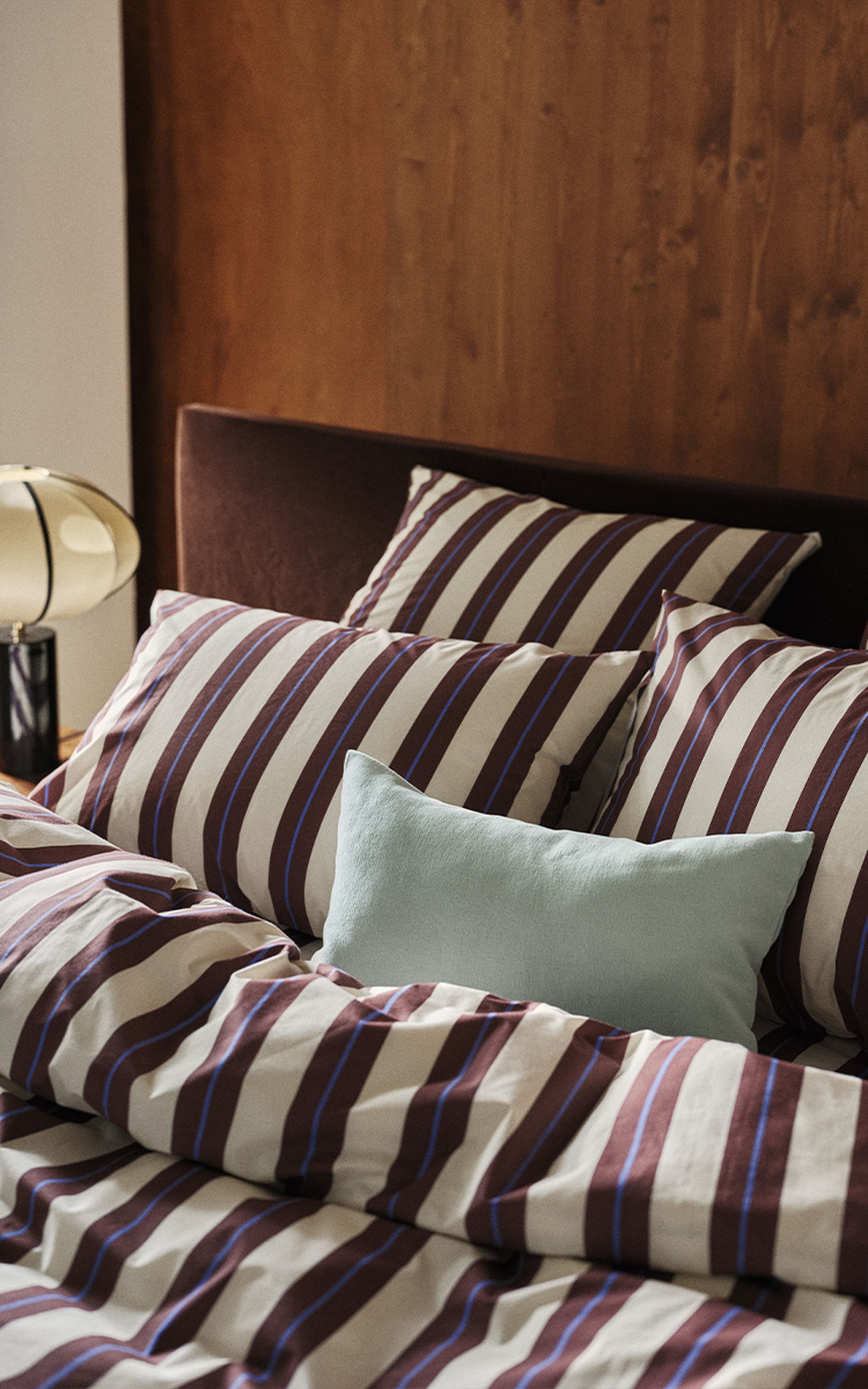 Un lit recouvert de draps rayés marron et blancs, avec plusieurs oreillers, dont un bleu, et une tête de lit en bois. On aperçoit partiellement une lampe sur le côté.