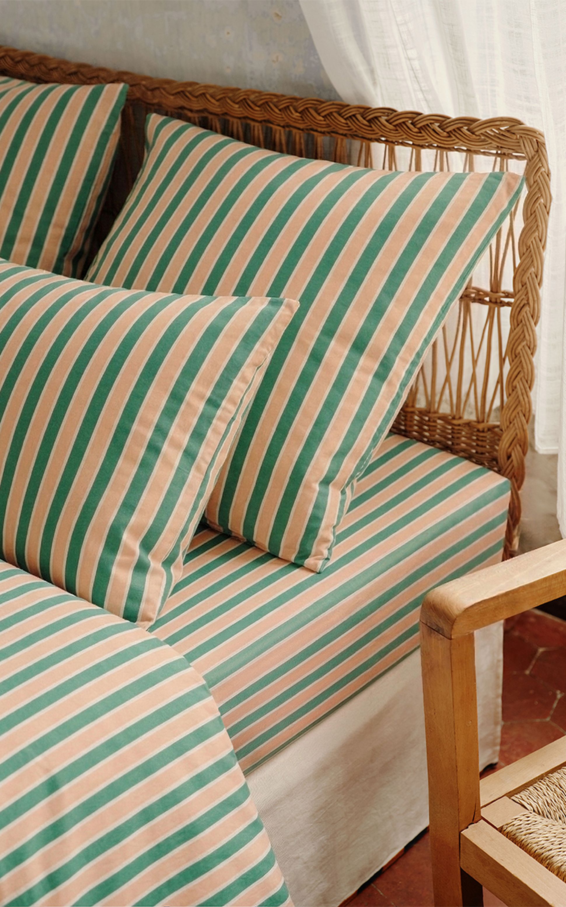 Un lit de repos en rotin avec des coussins rayés verts et beiges, à côté d'un fauteuil en bois et de rideaux blancs, dans un cadre chaleureux.