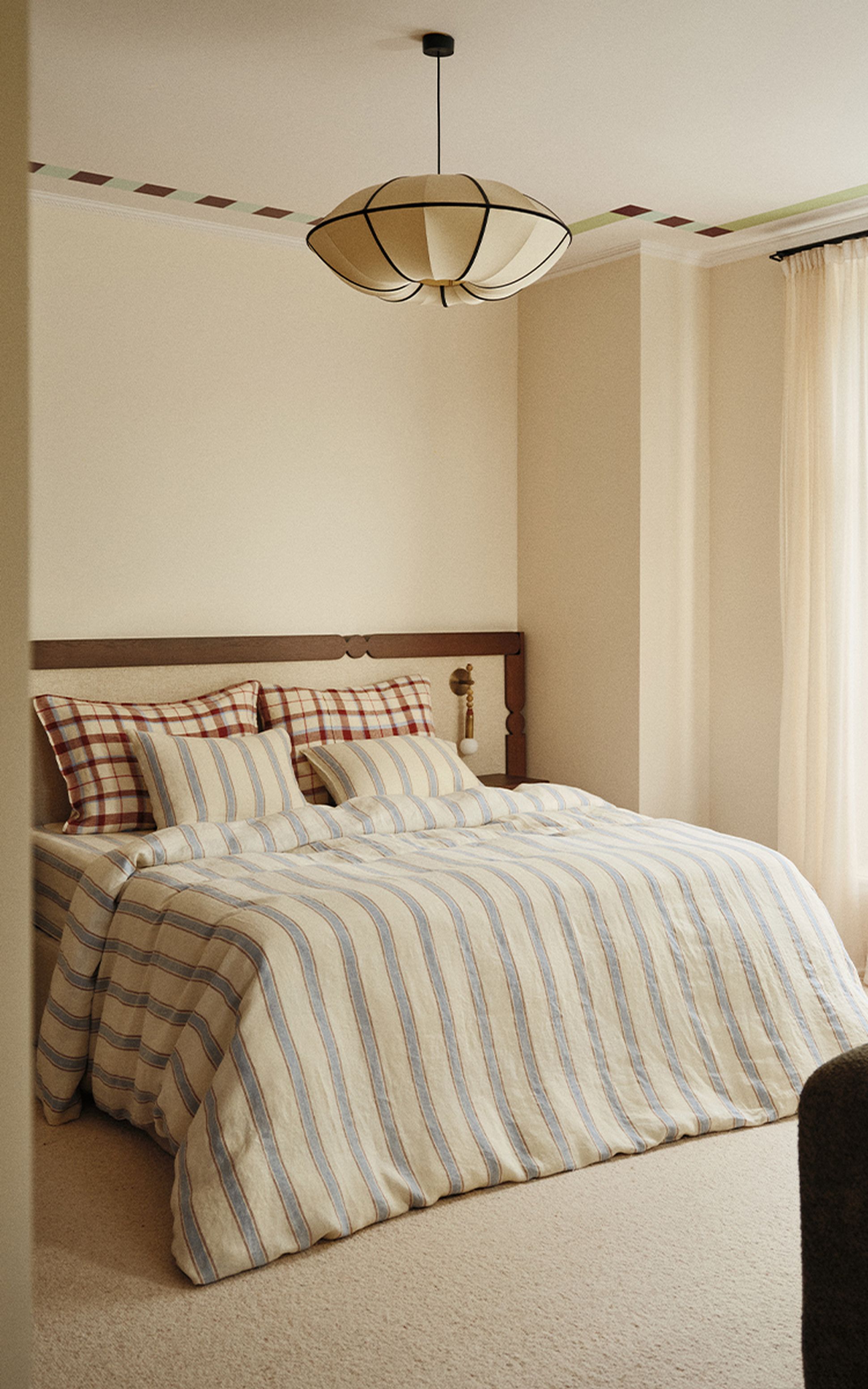 Chambre à coucher chaleureuse, avec un couvre-lit à carreaux et à rayures, des oreillers assortis et une tête de lit en bois. Une suspension est accrochée au plafond, et la pièce est agrémentée de rideaux de couleur claire.
