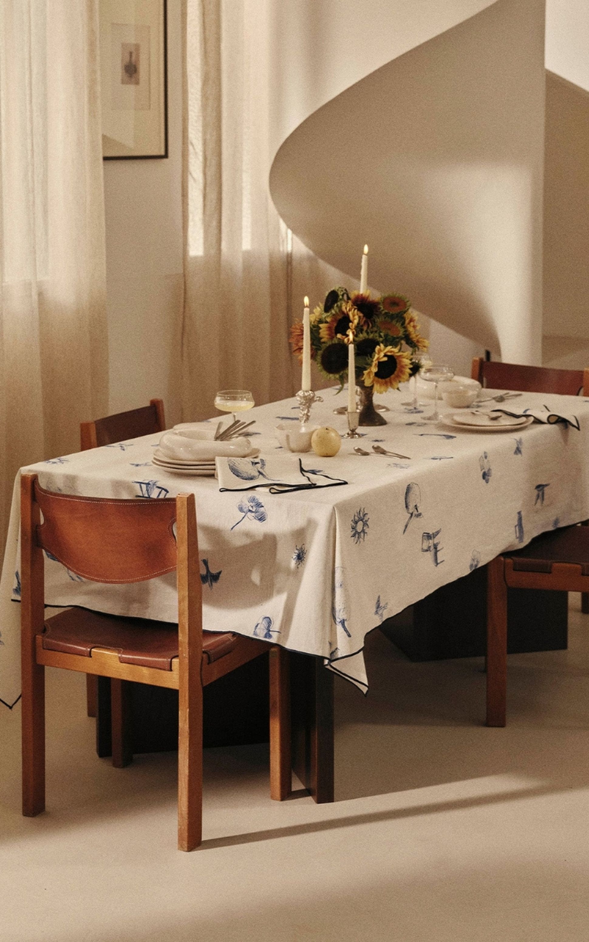 Une table à manger dressée avec une nappe imprimée, de la vaisselle élégante, des bougies, des tournesols et des chaises en bois, dans une pièce baignée d'une lumière tamisée.