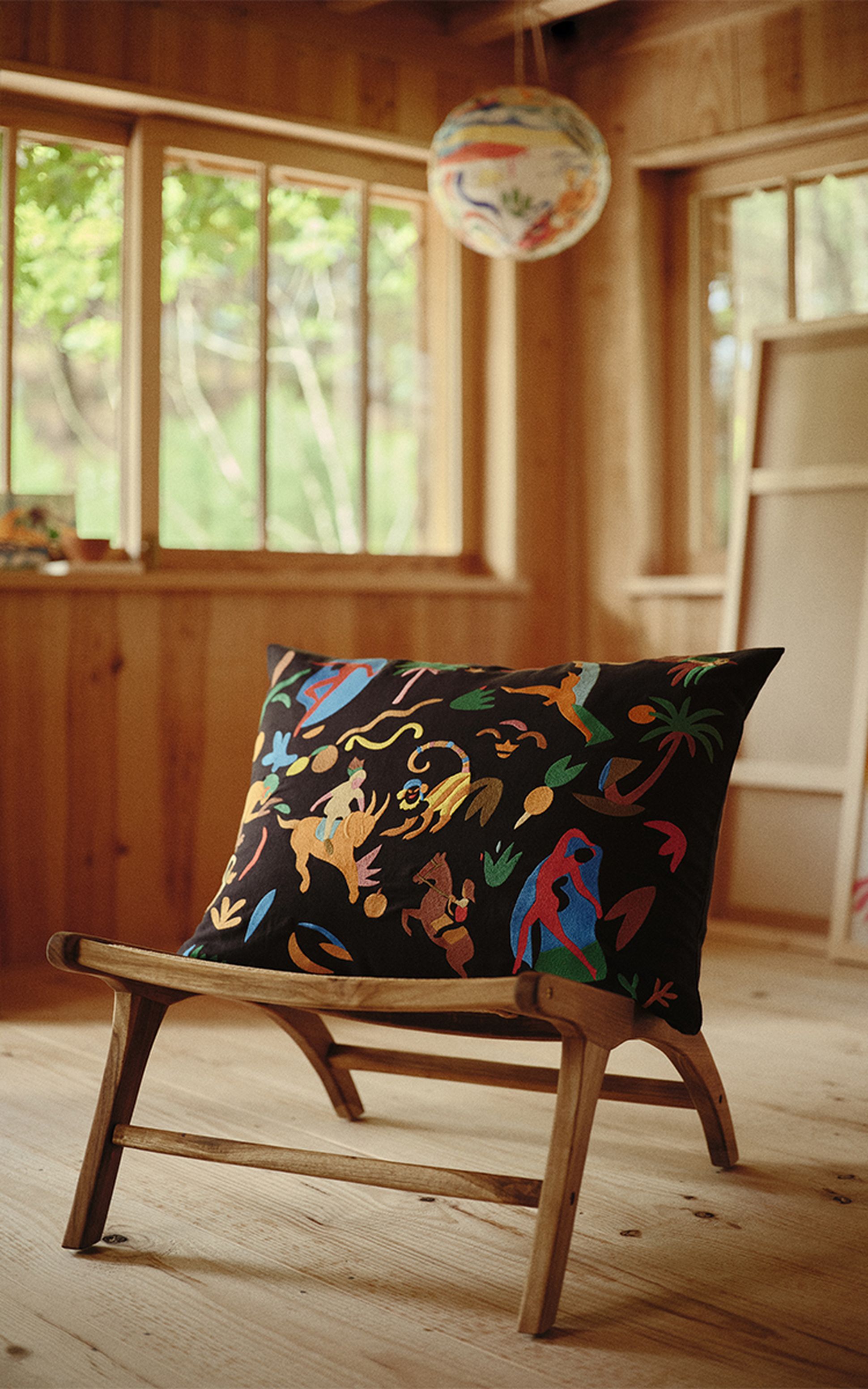 Un coussin coloré orné de motifs animaliers abstraits sur une chaise en bois, dans une pièce en bois baignée de soleil, dotée de grandes fenêtres et d'une lanterne en papier.