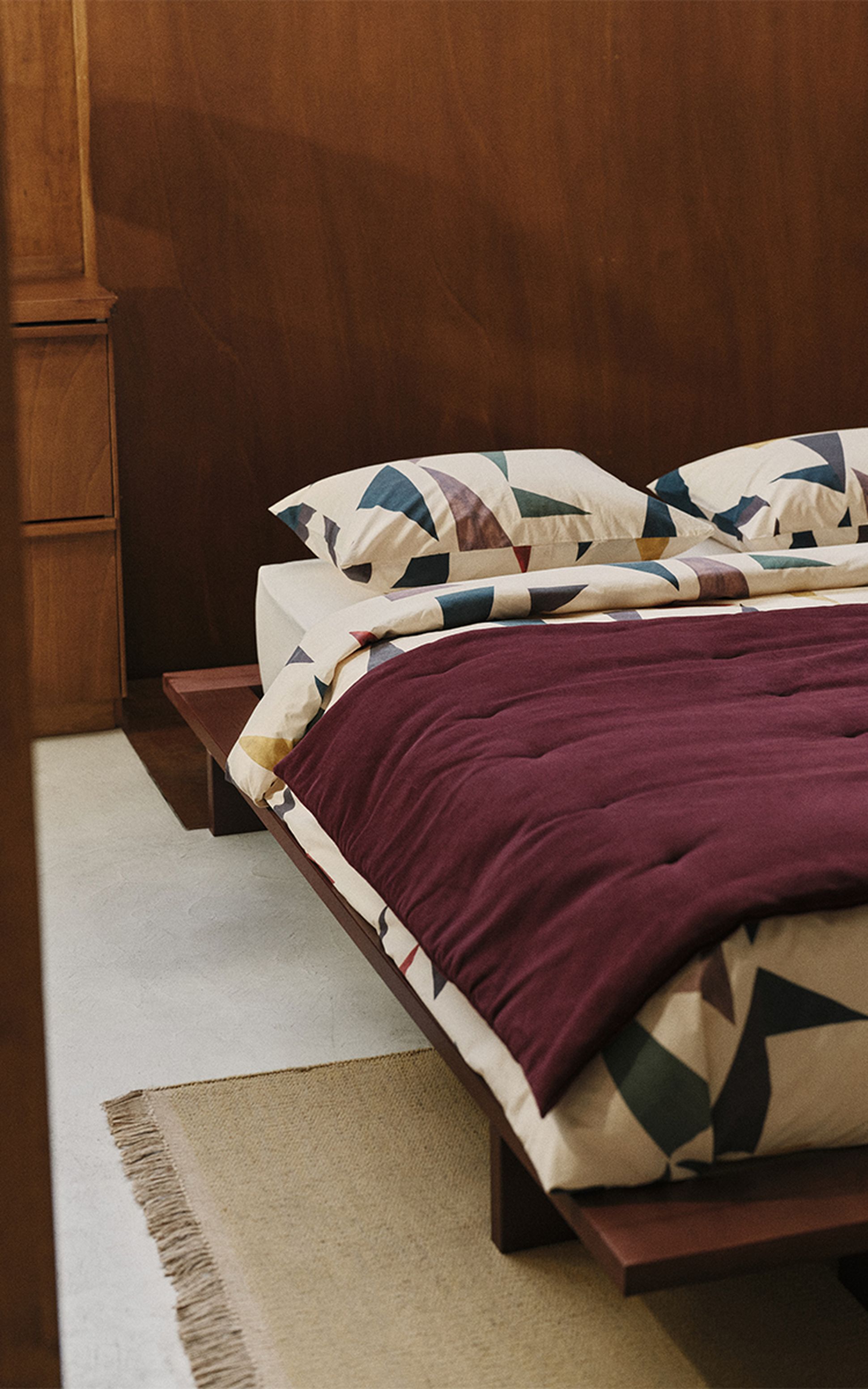 Un lit doté d'oreillers aux motifs géométriques et d'une couette bordeaux, posé sur un cadre en bois, dans une chambre aux murs en bois et agrémentée d'un tapis en jute.