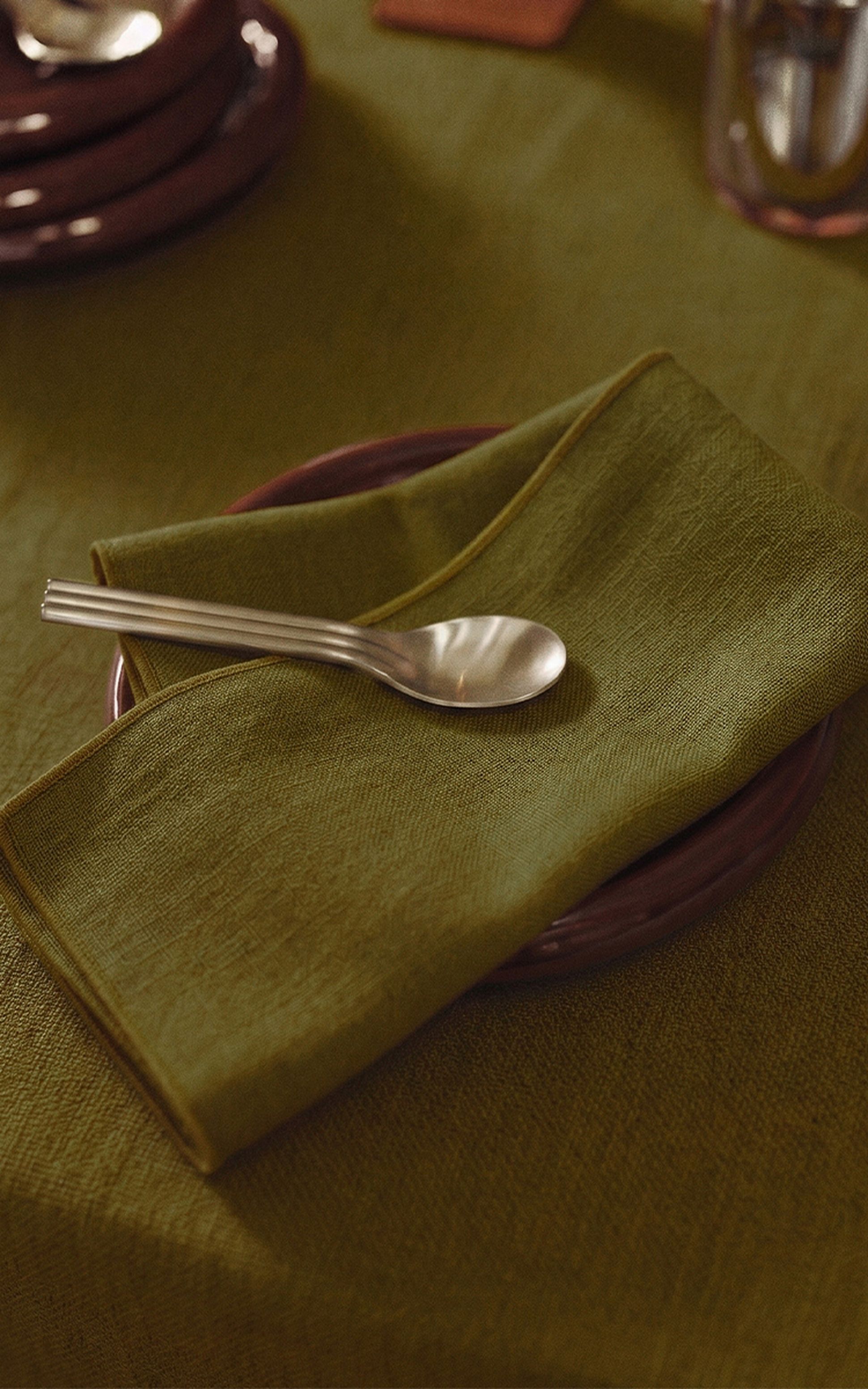 Une cuillère en argent repose sur une serviette en tissu verte posée sur une assiette marron vide, elle-même posée sur une nappe verte assortie.