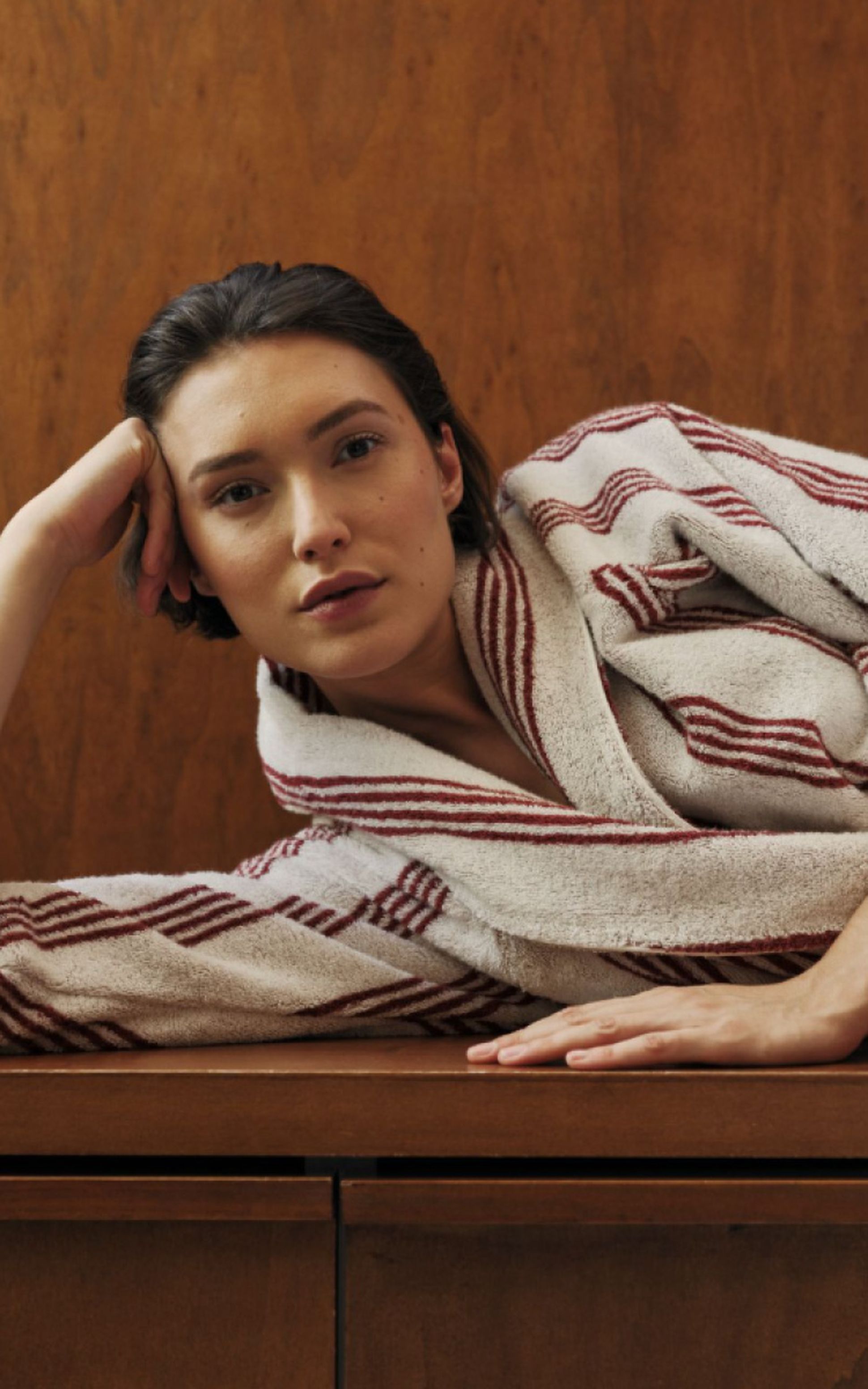 A person in a striped bathrobe leans sideways on a wooden surface, with one arm supporting their head.