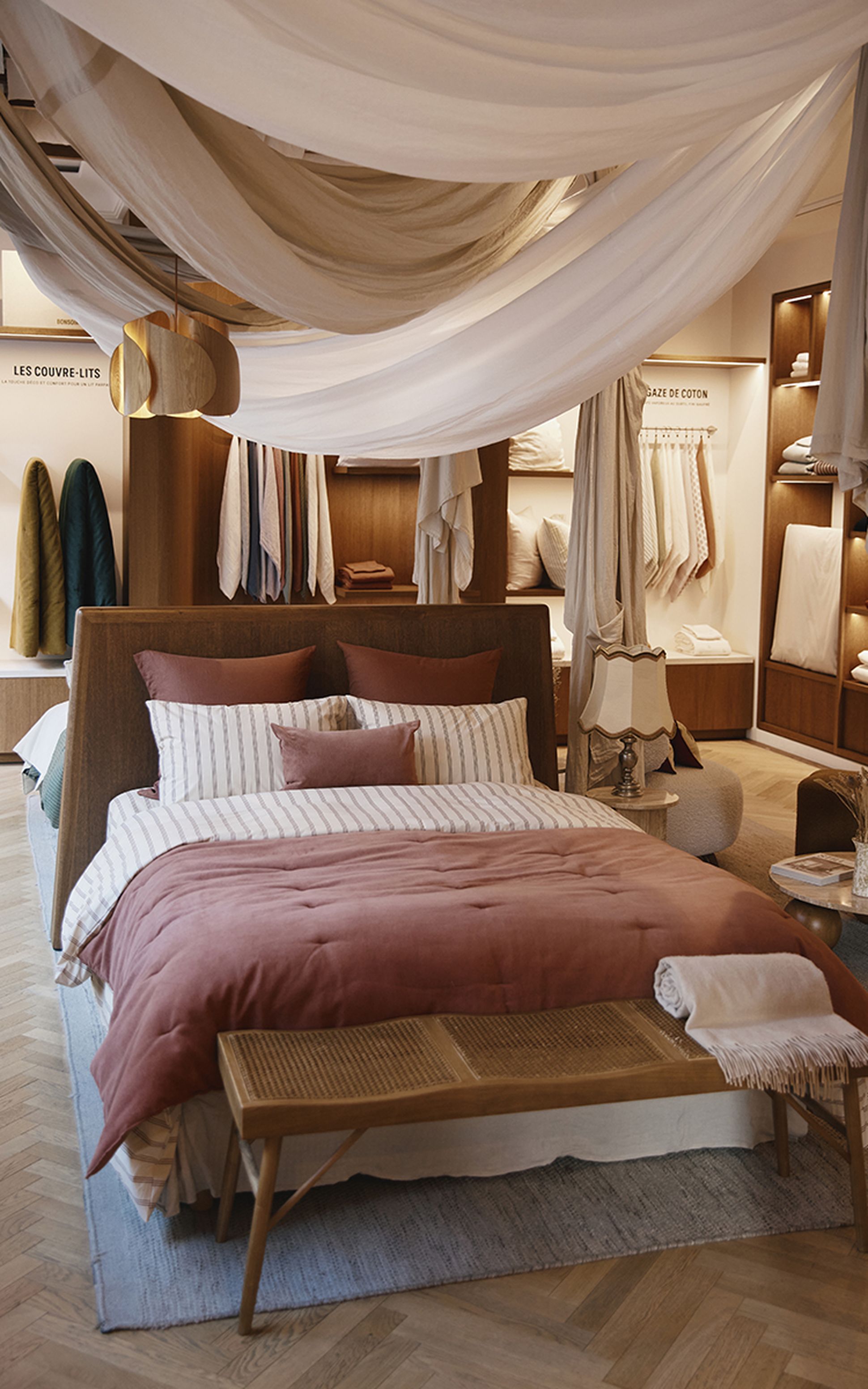 Une chambre à coucher chaleureuse, avec un lit habillé d'une parure à rayures et unise, entouré de tissus drapés et d'étagères en bois sur lesquelles sont disposés des linge de maison.